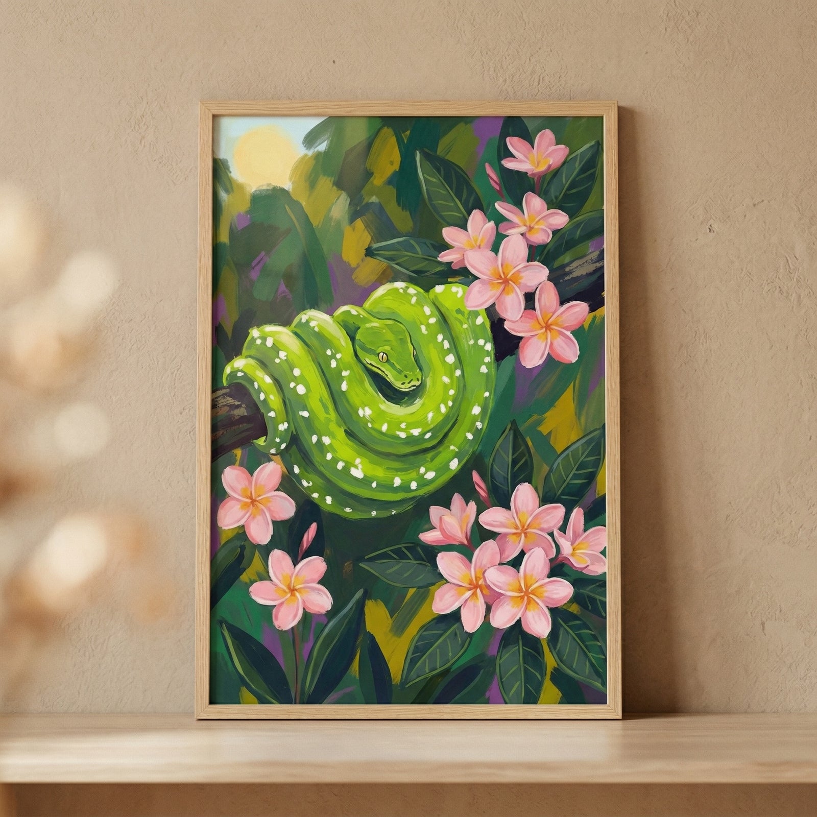vA vibrant green serpent with white speckles is depicted coiled on a dark branch in this stylized illustration. It is surrounded by lush tropical foliage and several pink blossoms with yellow-tinged centers. The background shows a soft, glowing yellow sun through abstract green and yellow leaves. The entire image is presented in a light-colored wood frame on a shelf against a neutral background.
