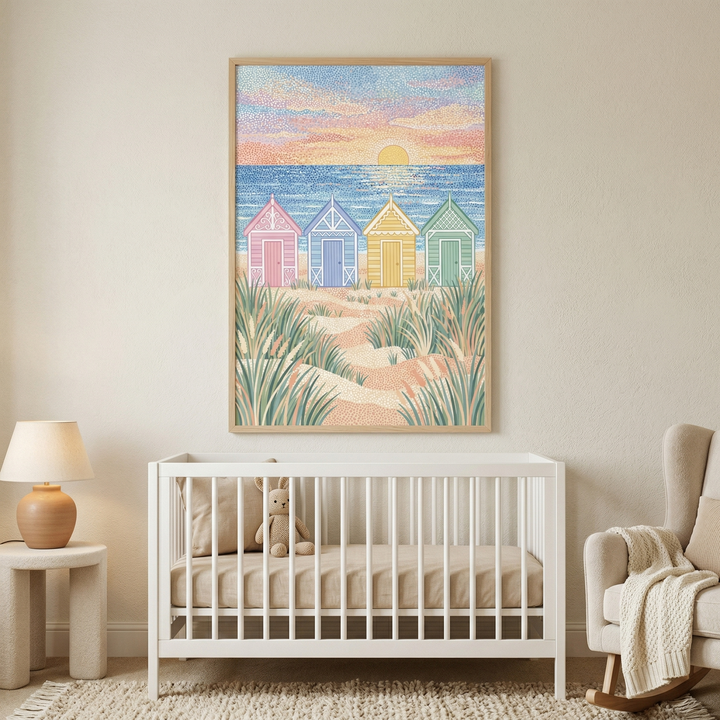 A bright and airy nursery features a large, framed piece of art hanging on a neutral-toned wall above a white wooden crib. The illustration within the frame depicts a peaceful coastal scene in a stippled art style. Four charming beach huts, colored in shades of soft pink, light blue, pale yellow, and mint green, stand along a sandy path lined with tall beach grass. Beyond the dunes, a calm sea meets a vibrant sky filled with dots of pink, orange, and blue, with a large, golden sun setting on the horizon.