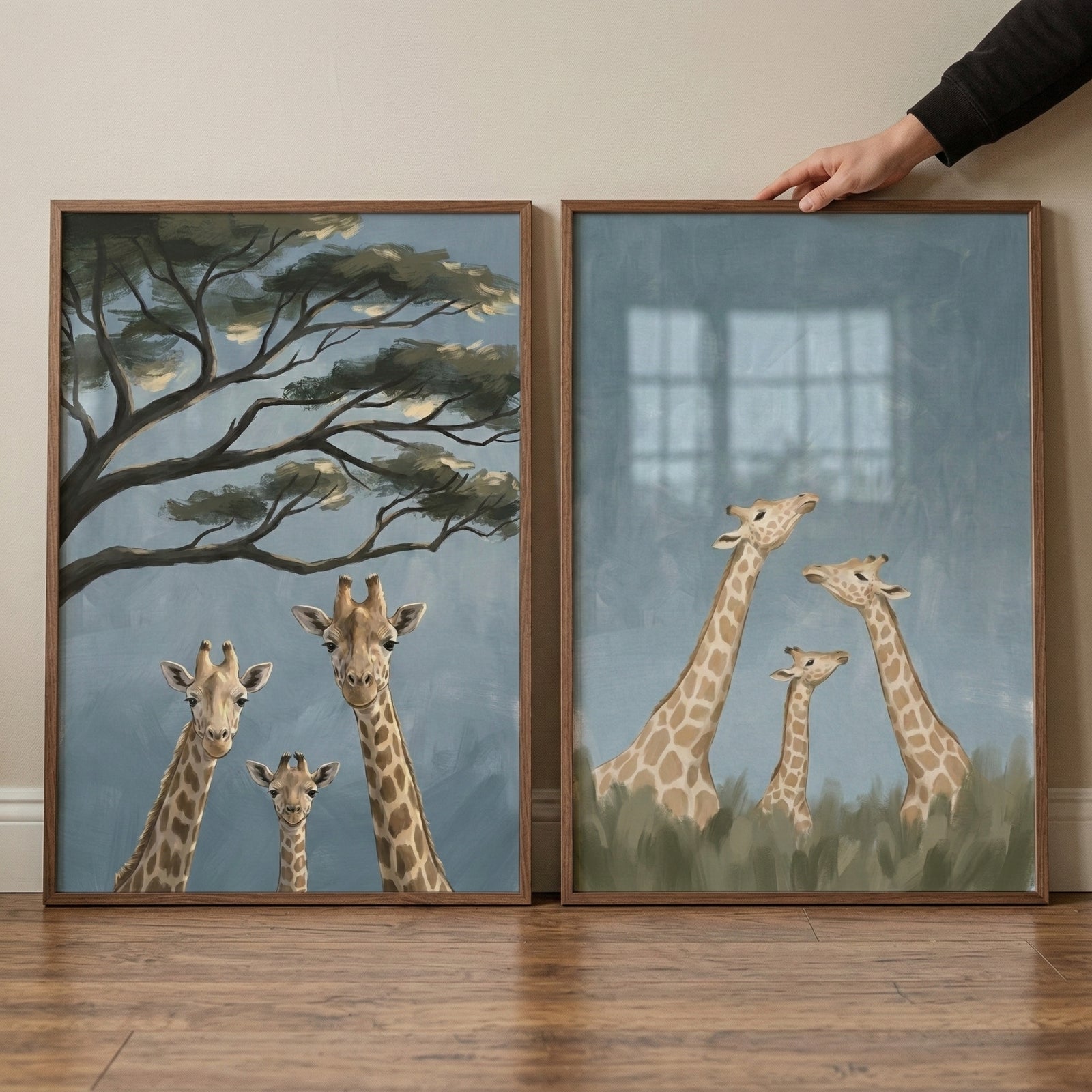Two framed giraffe paintings rest on a wooden floor against a cream wall. One artwork shows three giraffes looking forward beneath wide tree branches, while the other shows three giraffes rising above soft green grass against a muted blue background, with a hand holding the top corner to show scale.
