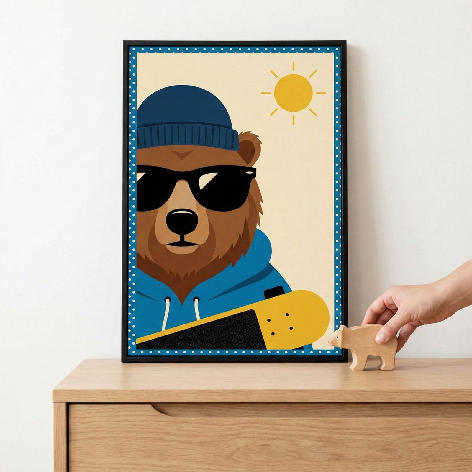 A framed illustration of a cartoon brown bear wearing a blue beanie and dark sunglasses, dressed in a blue hoodie. The bear holds a yellow and black skateboard under one arm against an off-white background featuring a simple yellow sun. The artwork has a blue and white polka-dot border and is housed in a thin black frame. The frame sits on a light-colored wooden dresser, and a person's hand is visible placing a small, plain wooden bear figurine next to it.