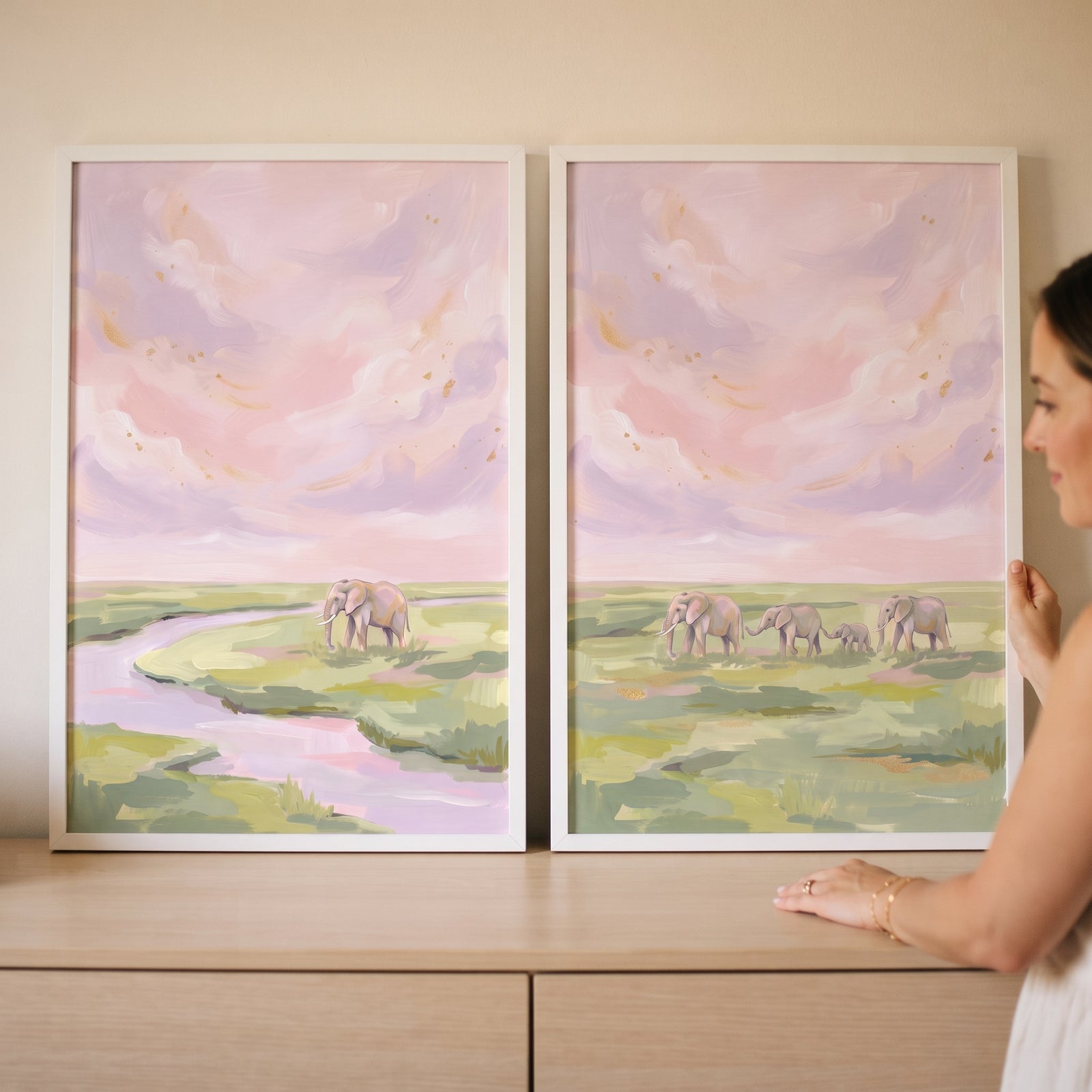 A woman holds a coquette blush pink elephant wall art set of 2 featuring elephants by a river in a pastel savanna.