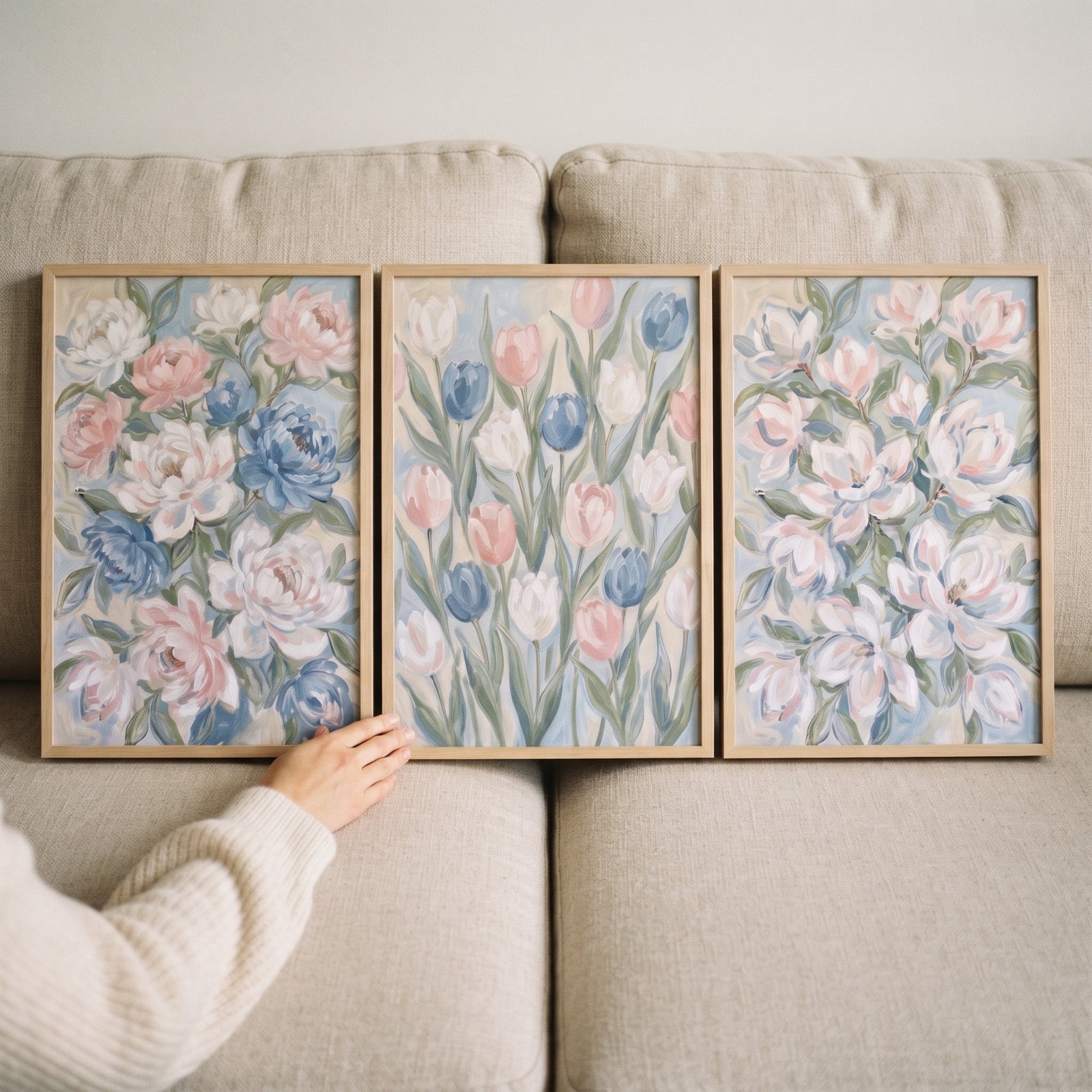 A hand touches a coquette pastel blue floral wall art set of 3 featuring pink and blue flowers on a light beige couch.