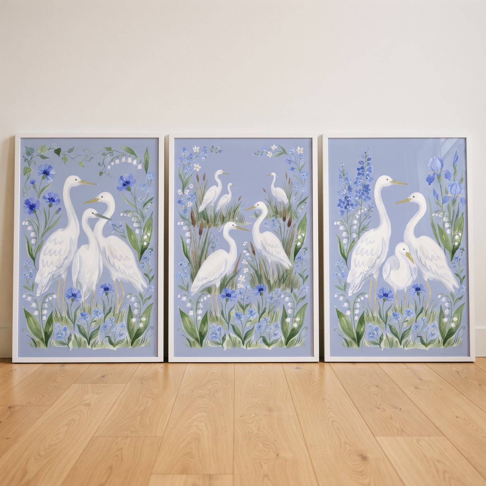 Cornflower blue botanical egret bird wall art set of 3 features white birds and blue flowers in frames on wood floors.