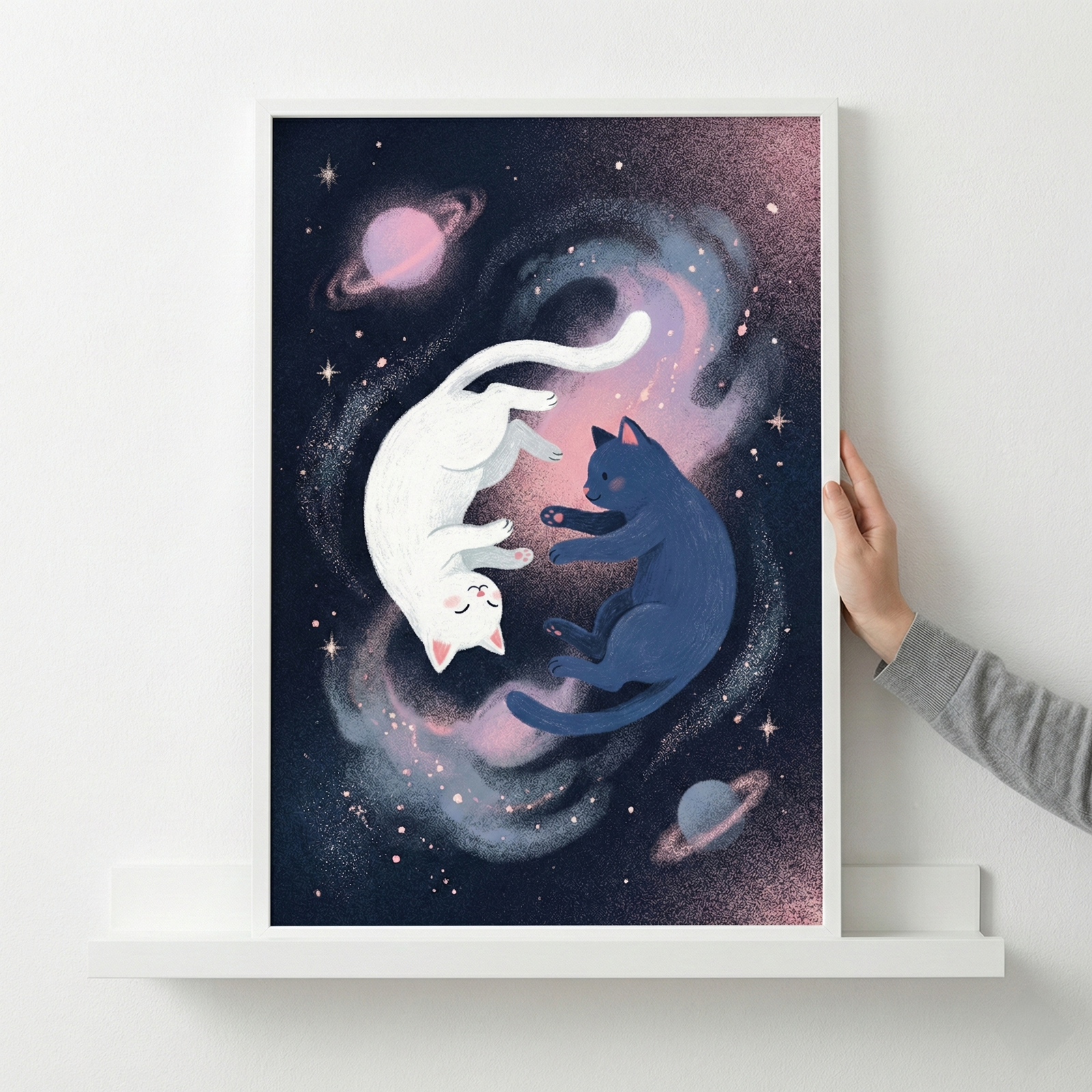 A framed cosmic illustration featuring two cats floating in space. One cat is white and the other is dark blue, positioned together in a circular shape. The background is a dark sky filled with soft, glowing pink and blue nebulae, small stars, and two ringed planets. The artwork is held by a hand on its right side as it rests on a white shelf against a white wall.