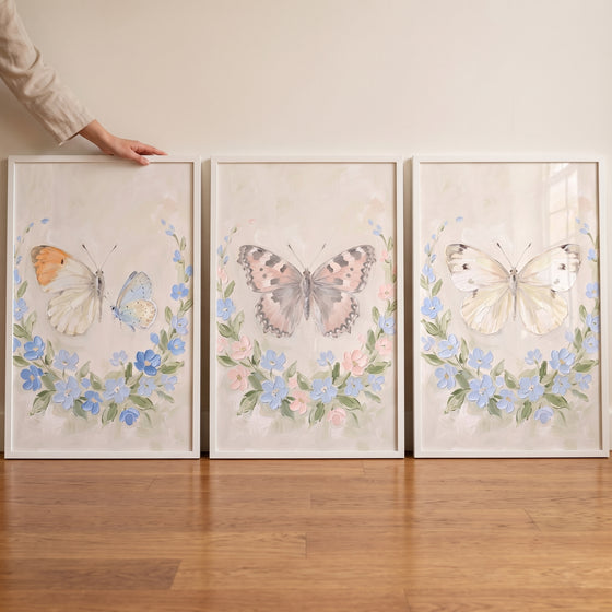 A hand touches a cottagecore floral wreath butterfly wall art set of 3 with soft blue and pink flowers on a wood floor.