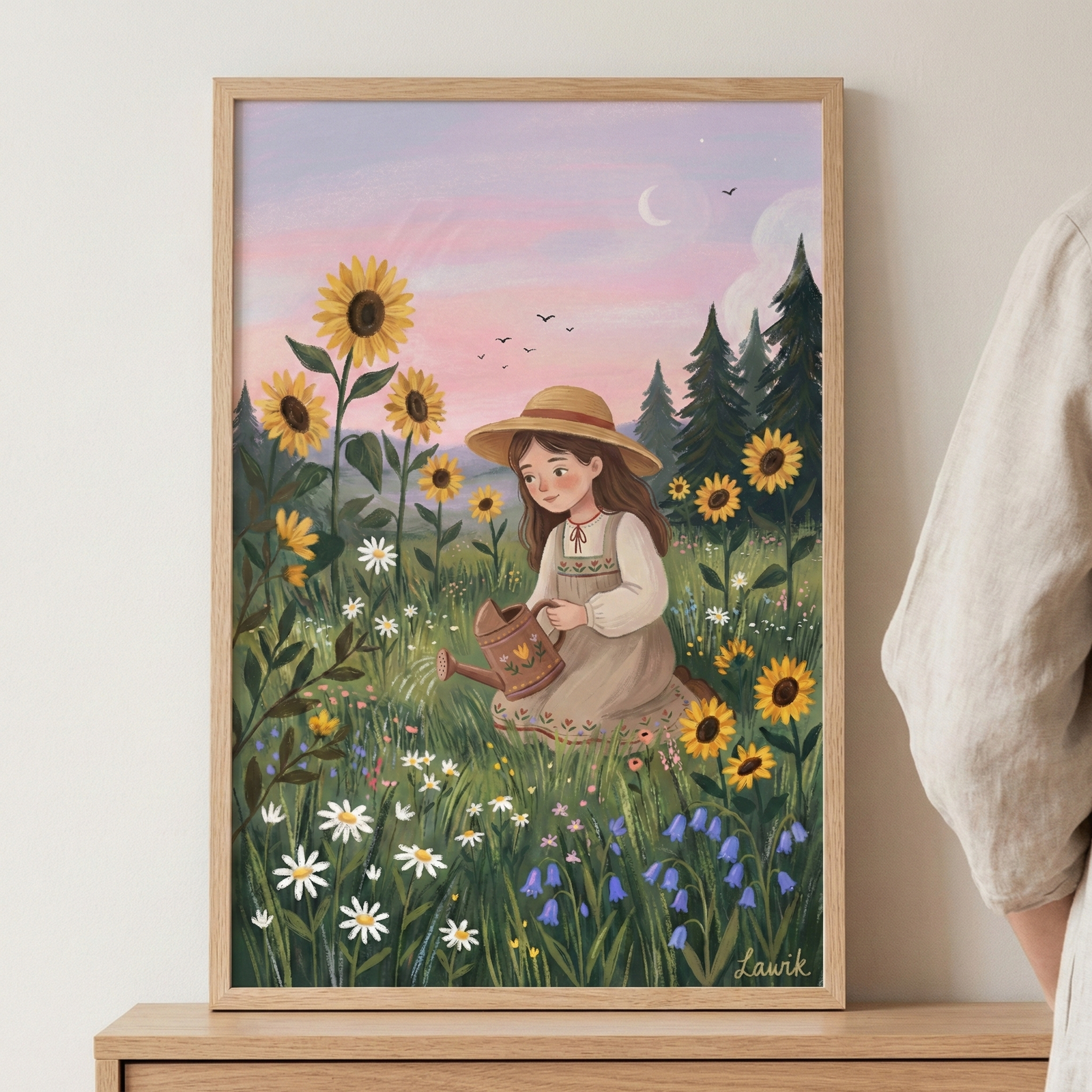 A vertical wood-framed illustration depicts a young girl with long brown hair and a straw hat kneeling in a lush field. She wears a white blouse under a tan pinafore dress and holds a small brown watering can. The field is vibrant with tall sunflowers, white daisies, and clusters of blue and purple wildflowers. Behind her are dark evergreen trees set against a soft pastel pink and lavender sky with a crescent moon and a small flock of birds flying in the distance.