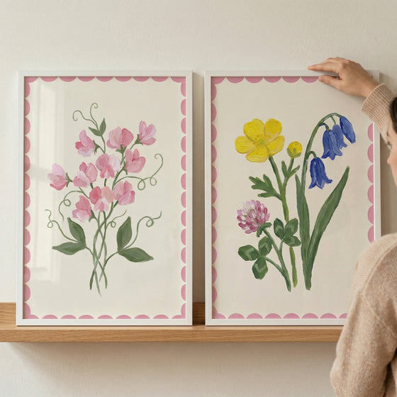 Two framed floral prints on a wooden shelf featuring soft pink sweet pea blossoms on the left and a mix of yellow blue and pink wildflowers on the right while a person adjusts the frame creating a styled home decor moment.