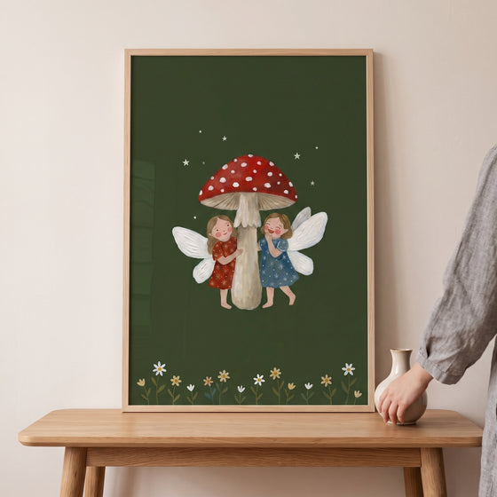 Cottagecore red toadstool fairies wall art with two fairies hugging a mushroom, framed on a wooden table.