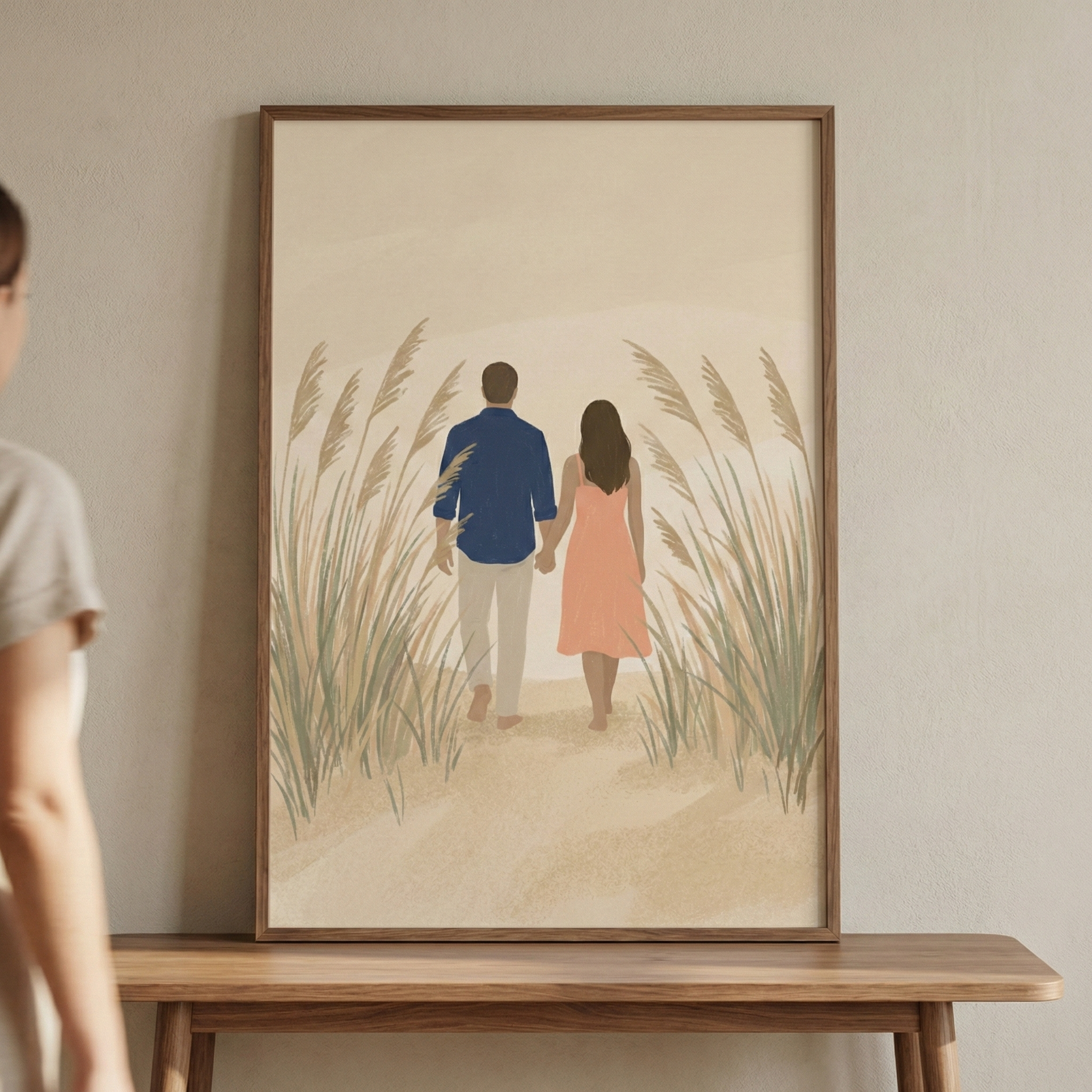 A minimalist, flat-style illustration captures a couple from behind, walking hand-in-hand along a sandy path. The man wears a navy blue shirt and light beige trousers, while the woman is in a soft peach-colored dress. They are surrounded by tall, feathery stalks of beach grass under a neutral, warm-toned sky. This artwork is displayed in a slender wooden frame, resting on a wooden table against a plain, light-colored wall.