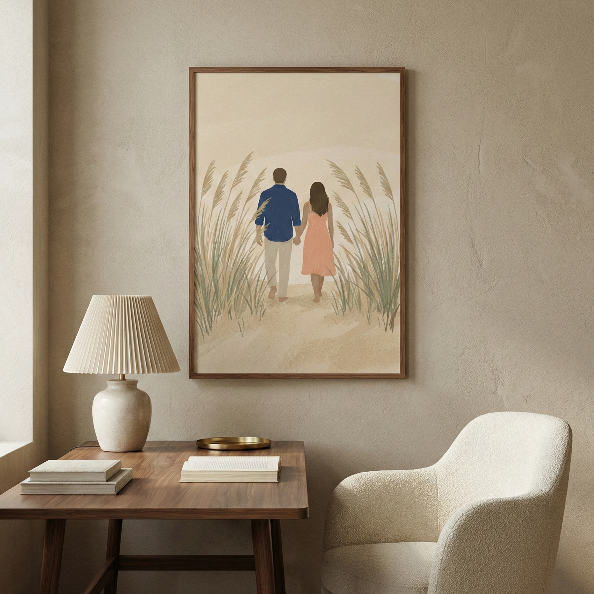 A minimalist framed art piece hangs on a light beige wall above a dark wood desk. The illustration depicts a man in a blue shirt and a woman in a peach dress walking away through tall grasses on a sandy path. On the desk, a cream-colored lamp with a pleated shade sits next to a stack of books and a small gold tray. To the right, a textured, off-white armchair completes the serene corner. The overall color palette is muted with earthy tones of tan, cream, and soft green.
