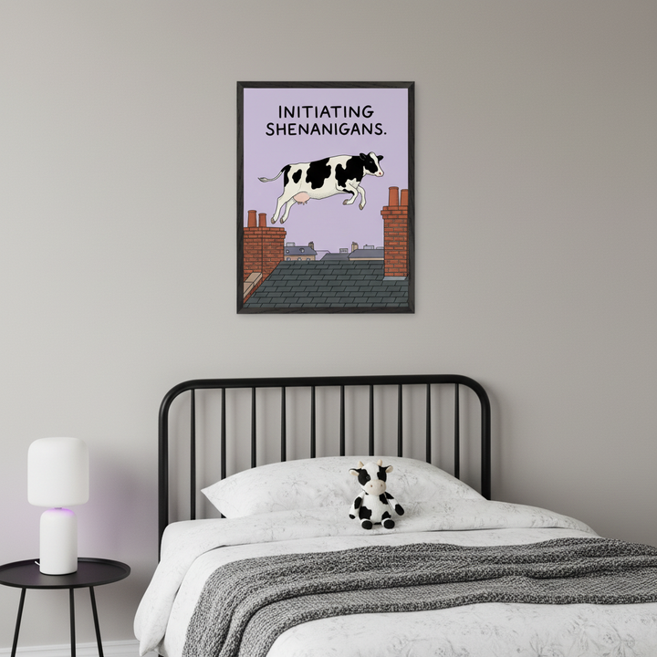 Modern kid's bedroom with a black metal bed, white bedding, gray throw, and cow plushie. A black side table with a white lamp is to the left. Above the bed, a framed print shows a cartoon black and white cow jumping over chimneys on a purple background with text: 'INITIATING SHENANIGANS'.