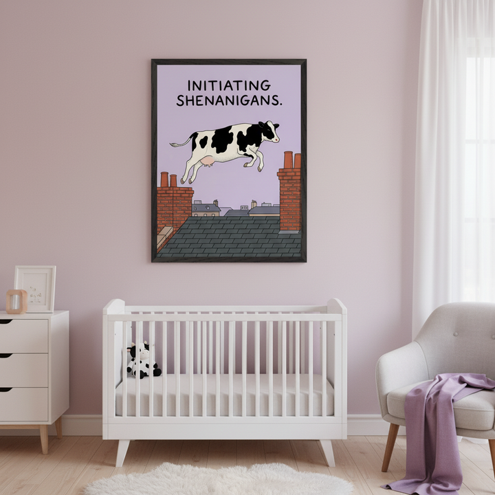 Modern nursery with a white crib, white dresser, and gray armchair. Above the crib, a large black-framed print depicts a cartoon black and white cow jumping over rooftops with "INITIATING SHENANIGANS" text against a purple sky.