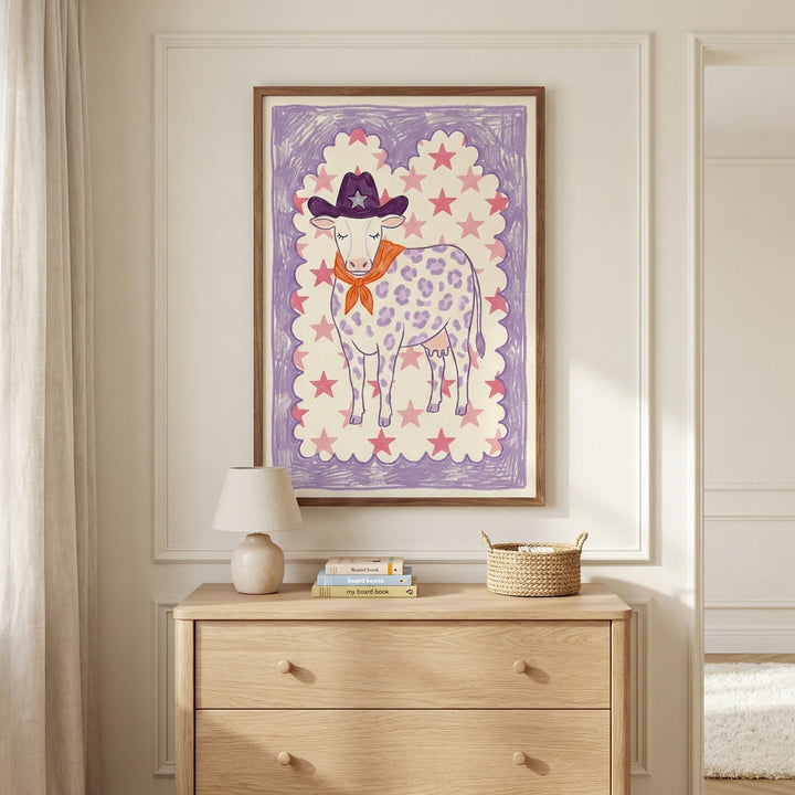 Framed western-themed cow artwork featuring star details and pastel tones, displayed above a wooden dresser in a softly lit, cozy nursery setting.