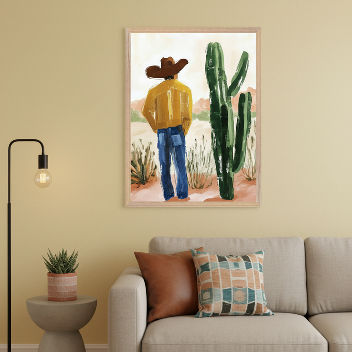 Modern living room with a beige sofa, throw pillows, and a floor lamp. A framed painting, showing a person in a cowboy hat observing a saguaro cactus in a desert, hangs above the sofa on a light yellow wall. A side table with a succulent sits below the lamp.