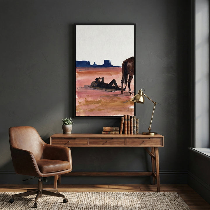 Western-themed art print of a cowboy lounging in the desert next to his horse, hung above a wood writing desk in a dark-toned study. The painting captures the stillness of the open range, with distant mesas and rugged textures conveying classic cowboy solitude.
