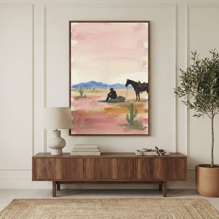 Framed watercolor painting of a cowboy sitting by a small campfire with smoke trailing into a soft pink sky, his horse standing nearby in a desert landscape with cacti and distant blue mountains. The artwork is displayed above a mid-century modern wood console in a neutral-toned room.