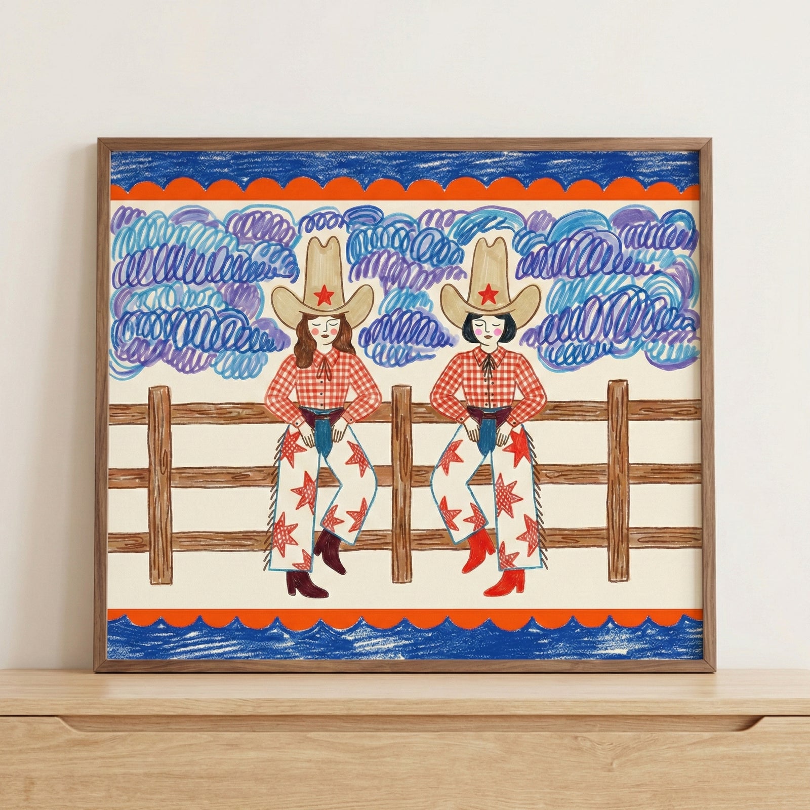 Close-up of framed cowgirl artwork featuring two illustrated women sitting on a wooden fence, with stylized purple clouds and a vibrant red and blue border. The hand-drawn western scene brings a colorful, nostalgic touch to modern wall art collections.