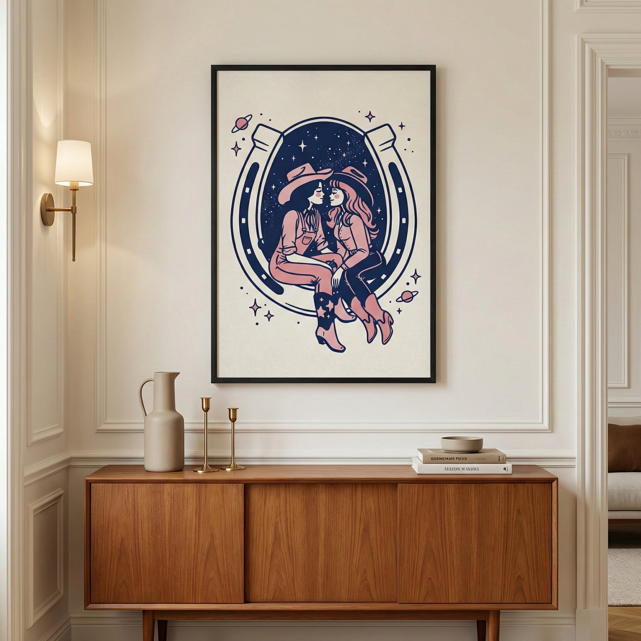Framed artwork featuring two cowgirls sitting close together inside a large horseshoe, set against a cosmic background with stars and planets, hangs above a mid-century modern wooden console. The room has elegant wall molding, warm lighting, and minimalist decor with ceramic vases and stacked books.
