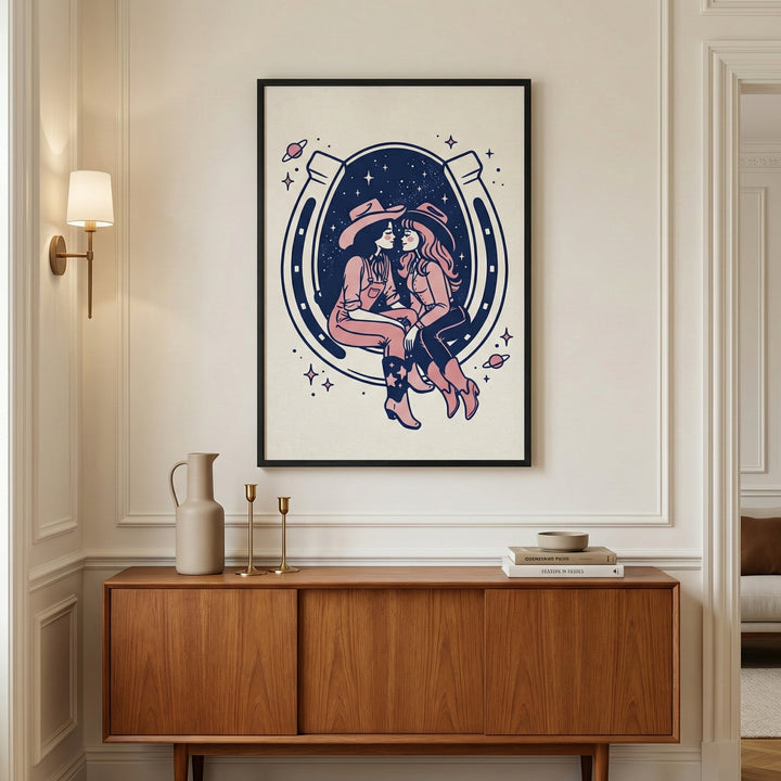 Framed artwork featuring two cowgirls sitting close together inside a large horseshoe, set against a cosmic background with stars and planets, hangs above a mid-century modern wooden console. The room has elegant wall molding, warm lighting, and minimalist decor with ceramic vases and stacked books.

