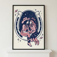 Framed illustration of two women dressed as cowgirls, sitting close and gazing at each other affectionately inside a large horseshoe. The background features a starry night sky with planets and sparkles, blending western and cosmic themes in a bold navy and pink color palette.
