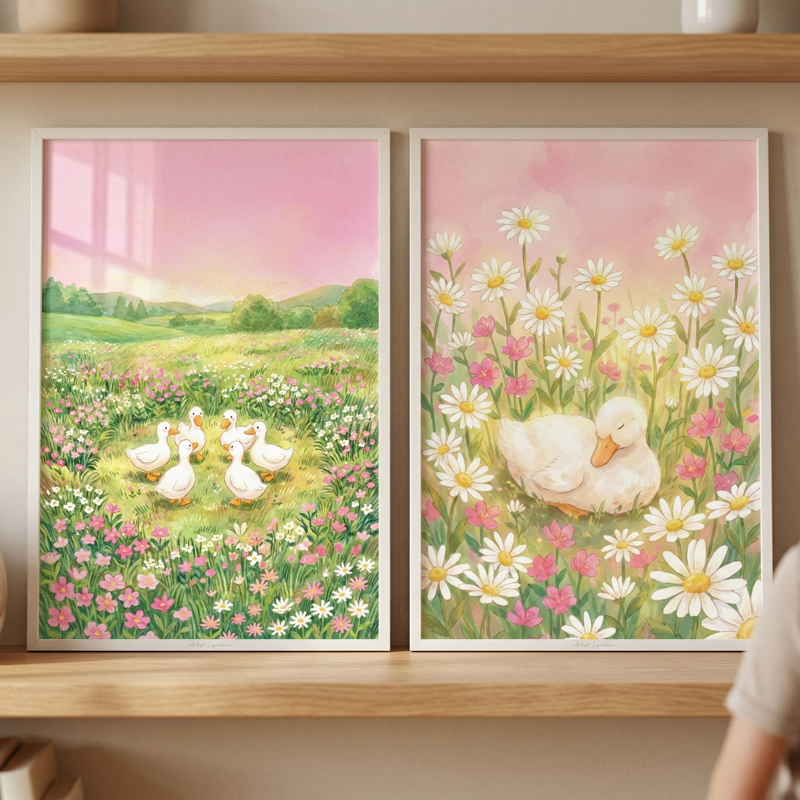Two white-framed illustrations rest on a light wood shelf against a neutral wall. The left artwork features a group of six small white ducklings in a lush, flower-filled meadow beneath a soft pink sky. The right artwork shows a close-up of a single white duckling nestled among large white daisies and small pink flowers, appearing to sleep against a pastel pink backdrop. Both scenes have a gentle, whimsical watercolor style with a warm, serene atmosphere.