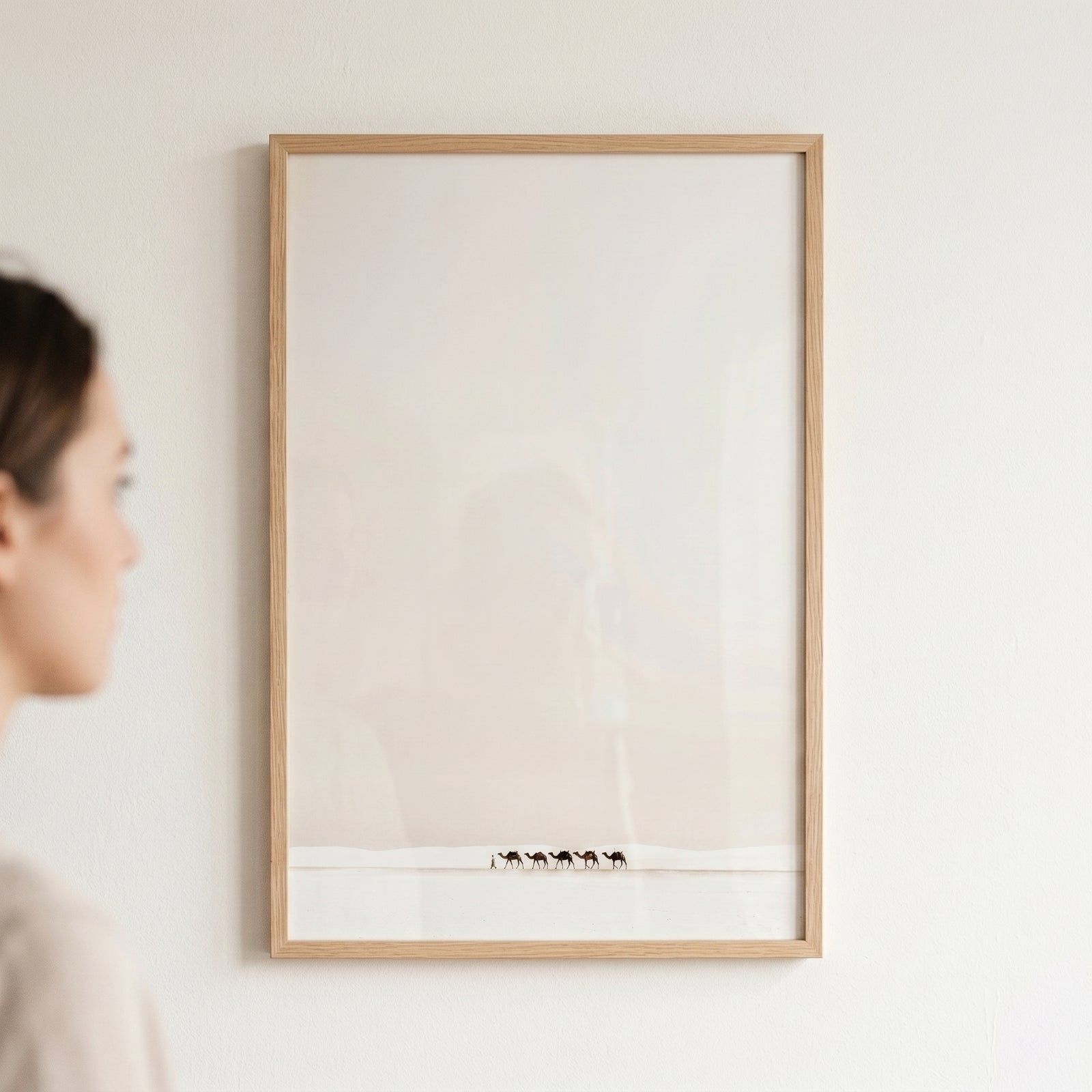 Framed creamy minimalist white camel wall art hangs on a white wall next to a person, showing a desert caravan.
