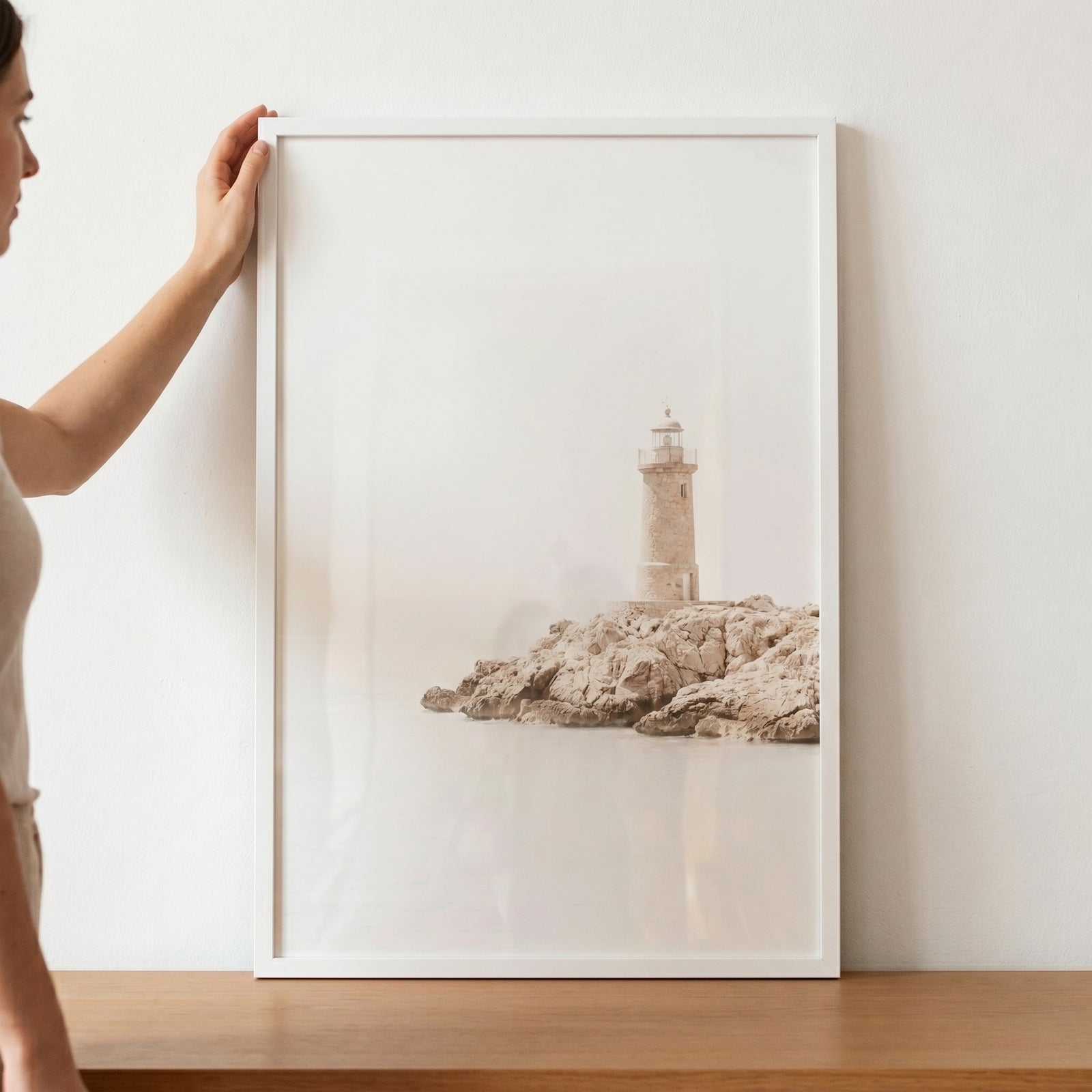 A person holds creamy neutral coastal lighthouse wall art in a white frame against a white wall above a wooden surface.