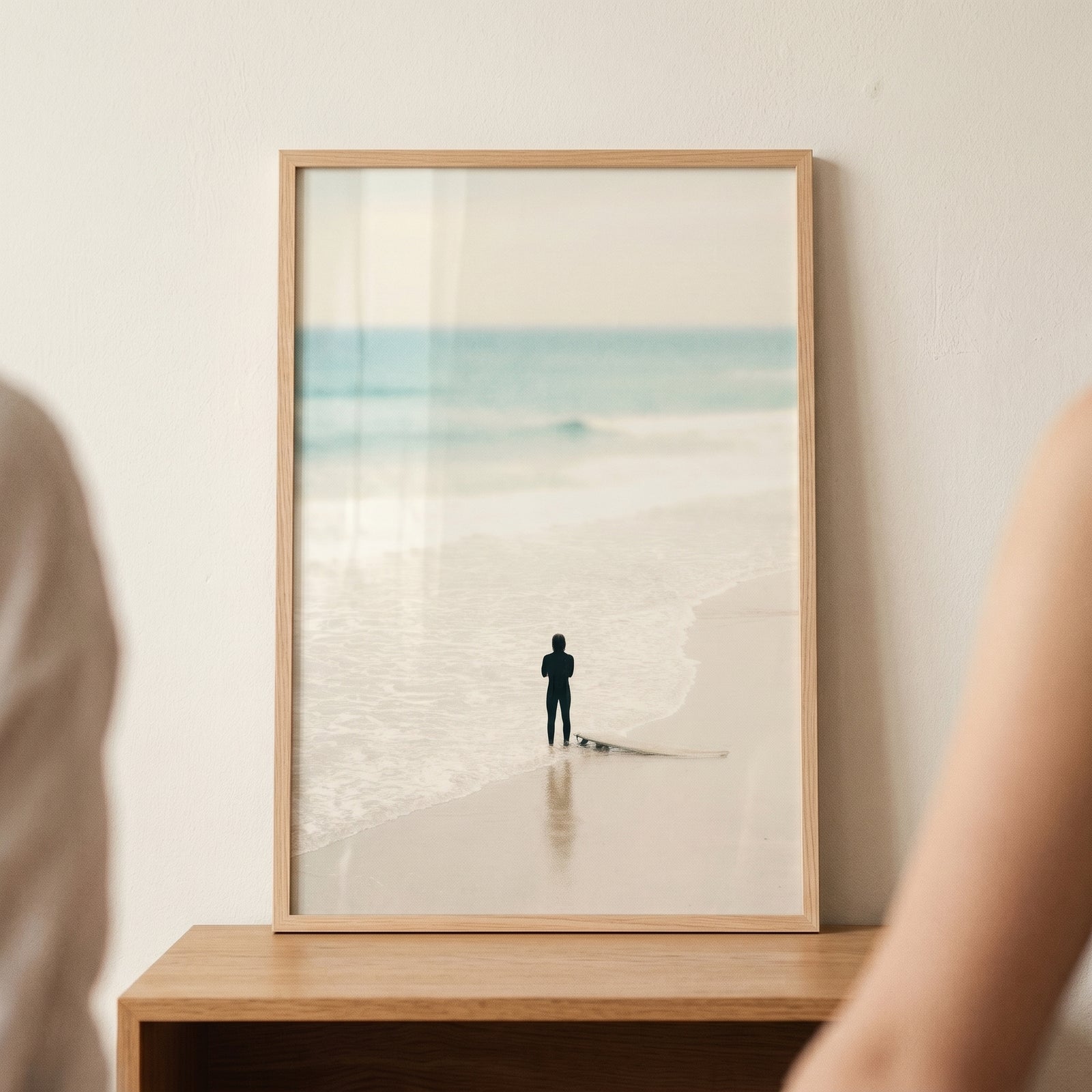 Framed creamy neutral minimalist surfer wall art with a lone figure and surfboard on a pale beach beside the ocean.