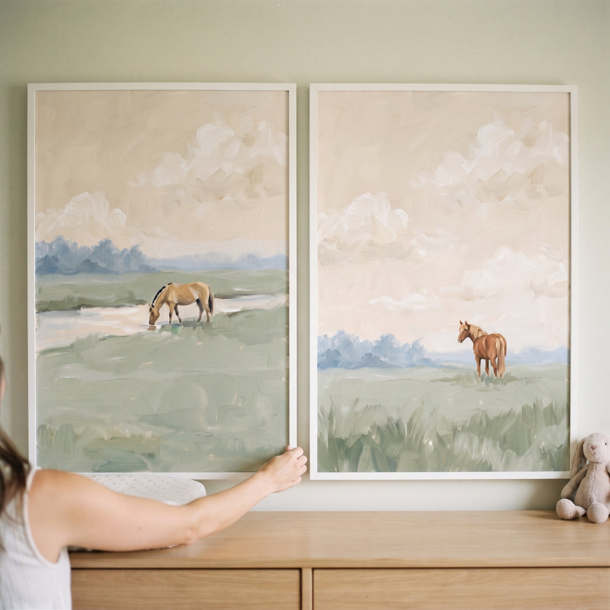Hand adjusting a creamy peach sky horse wall art set of 2 above a dresser with a plush rabbit nearby.