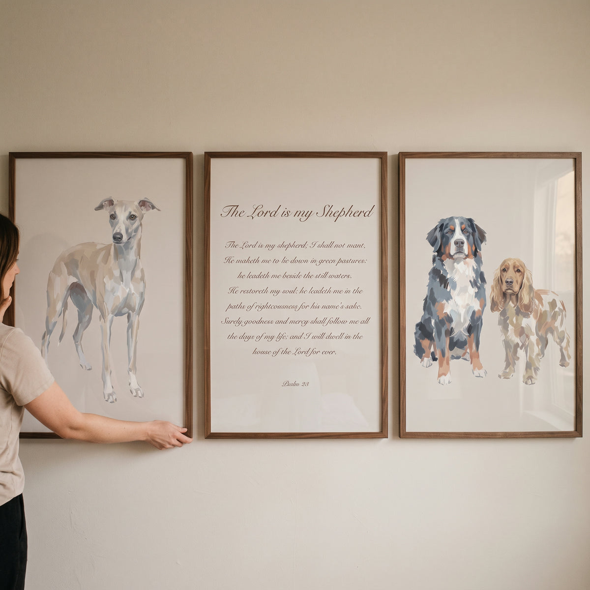 A person hangs a creamy sand scripture dogs wall art set of 3 featuring various dog breeds and Psalm 23 on a neutral wall.