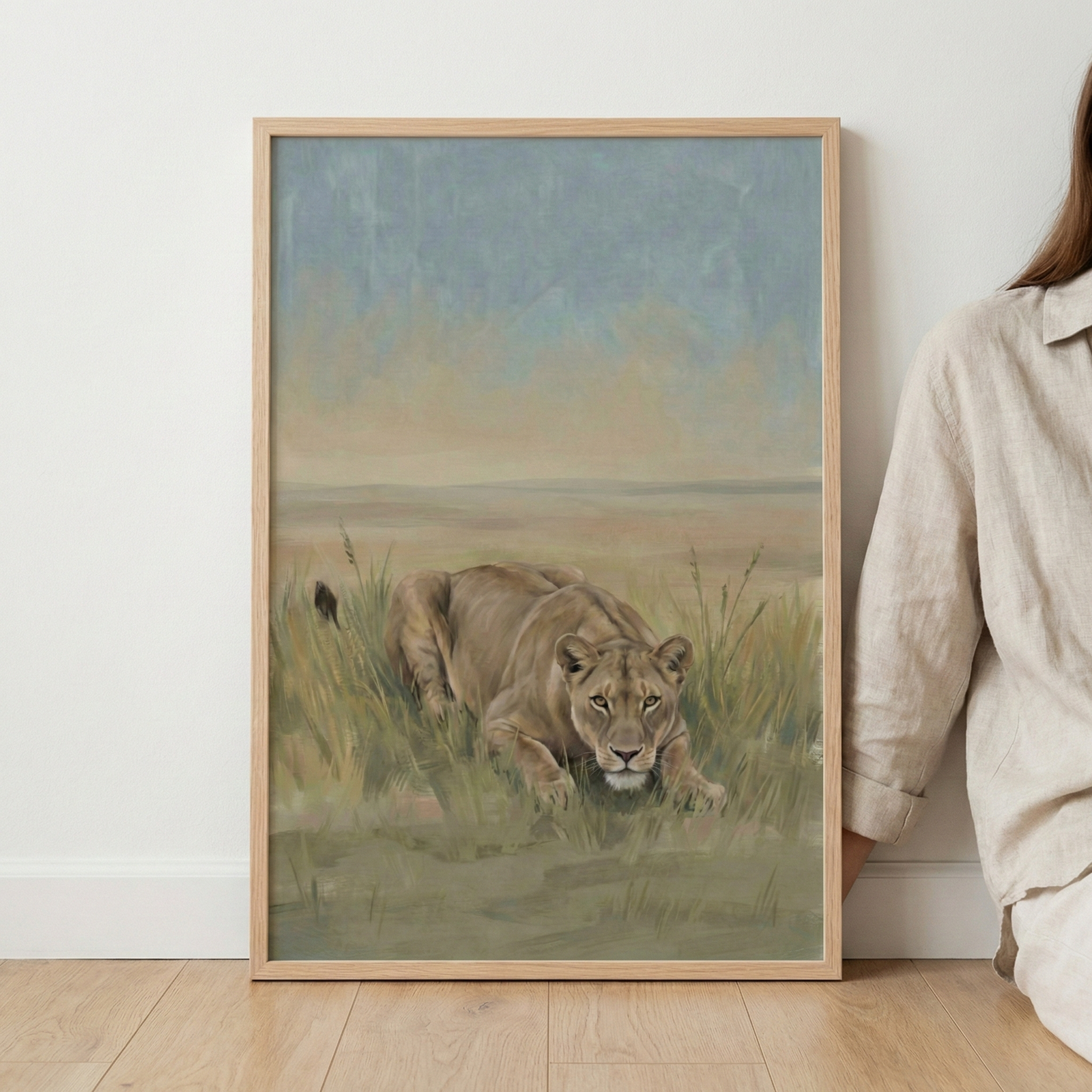 A framed piece of artwork featuring a lioness crouching in tall golden grass. The animal is depicted looking forward with a steady gaze. The background is a vast, open landscape under a soft sky of blue and tan tones. The frame is a light-colored wood and is leaning against a plain white wall on a light-colored wooden floor. Part of a person wearing a beige linen shirt is visible on the right.