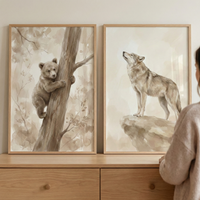 Two vertical wooden frames with watercolor-style art sit atop a light-toned wooden dresser. The left frame features an illustration of a brown bear cub climbing a tree and looking forward. The right frame shows a wolf standing on a rocky outcrop, its head tilted upward toward the sky. Both pieces are rendered in soft, neutral shades of beige and brown. A person in a light-colored sweater is partially visible on the right side as they look at the two pieces of art.