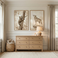 Two soft sepia wildlife illustrations hang above a light wood dresser in a cozy nursery. One artwork shows a bear cub climbing a tree trunk while the other shows a wolf standing on a rocky ledge with teddy bears and a woven basket of plush toys nearby.
