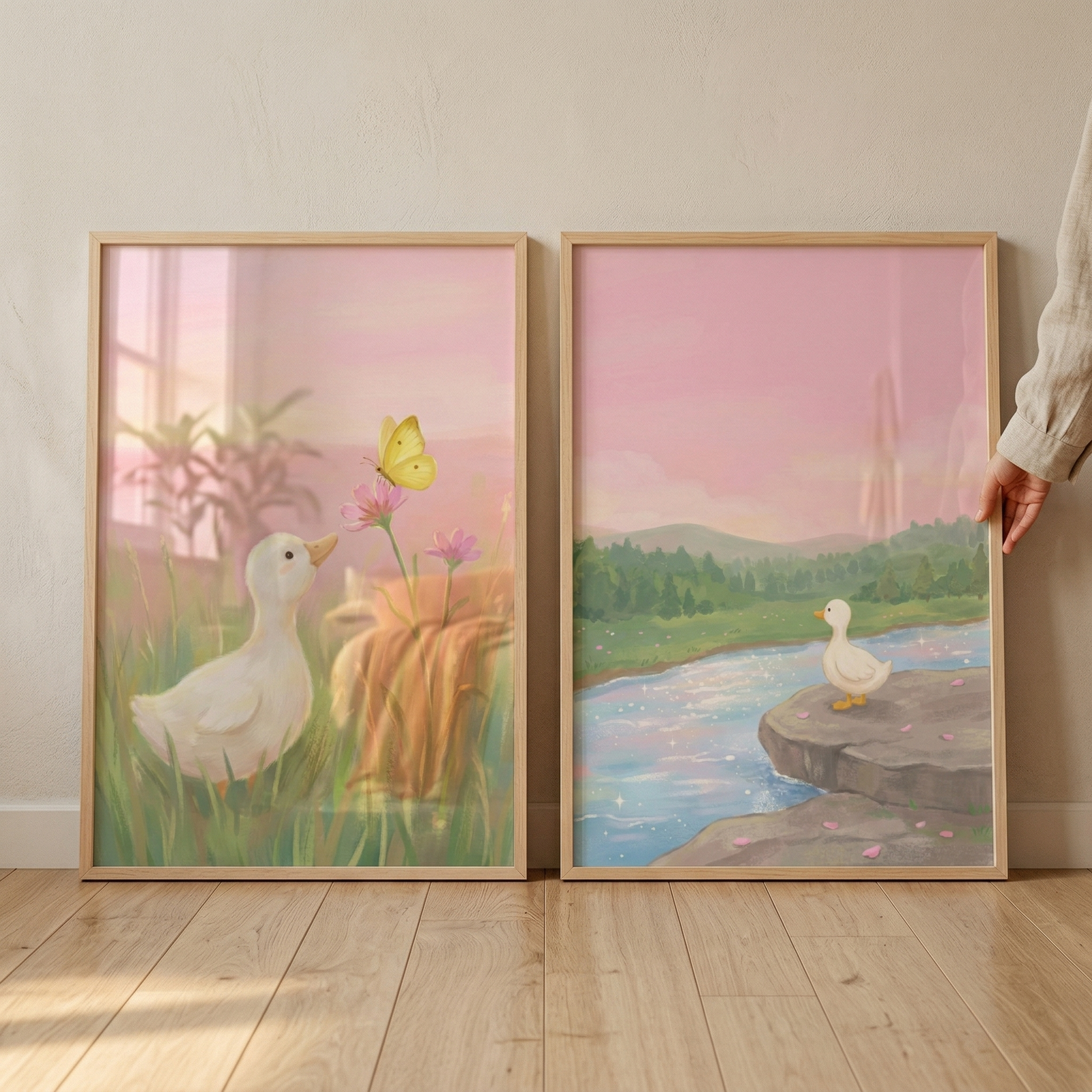 Two framed vertical rectangular pieces of art lean side-by-side against a plain, off-white wall on a light-colored wood floor. On the far right, a hand in a tan linen sleeve reaches out to steady the frame on the right. Both illustrations feature a soft, pastel color palette and a whimsical, storybook aesthetic.

The illustration on the left shows a close-up of a white duckling with a small yellow beak standing in tall green grass. It looks upward at a bright yellow butterfly resting on a tall pink flower.