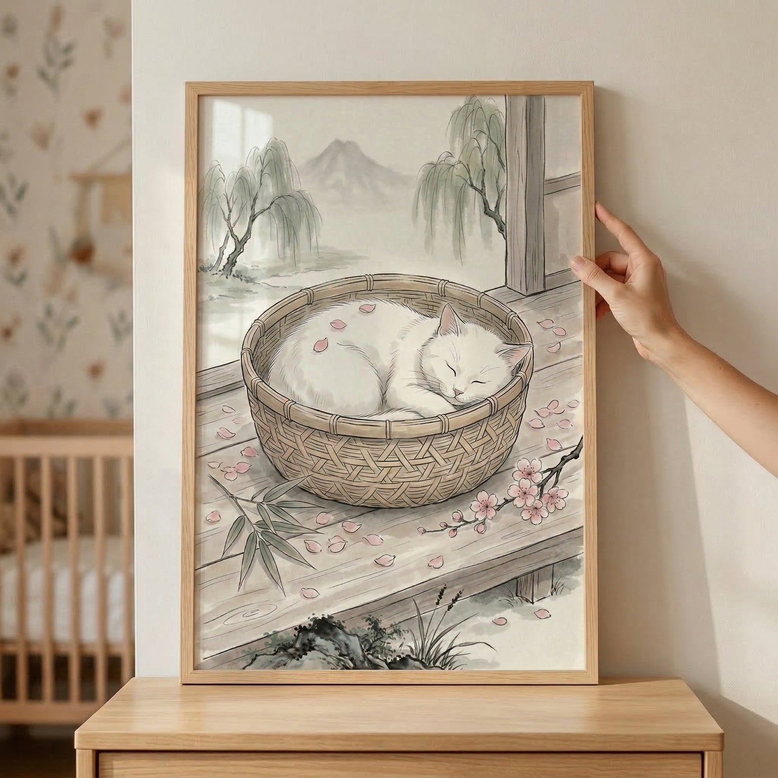 A framed illustration depicts a serene scene in a traditional East Asian ink wash style. Centered in the artwork is a peaceful white cat curled up and sleeping soundly inside a round, woven basket. The basket rests on a wooden porch floor where several delicate pink cherry blossom petals have fallen, some even resting on the cat's soft fur. 
