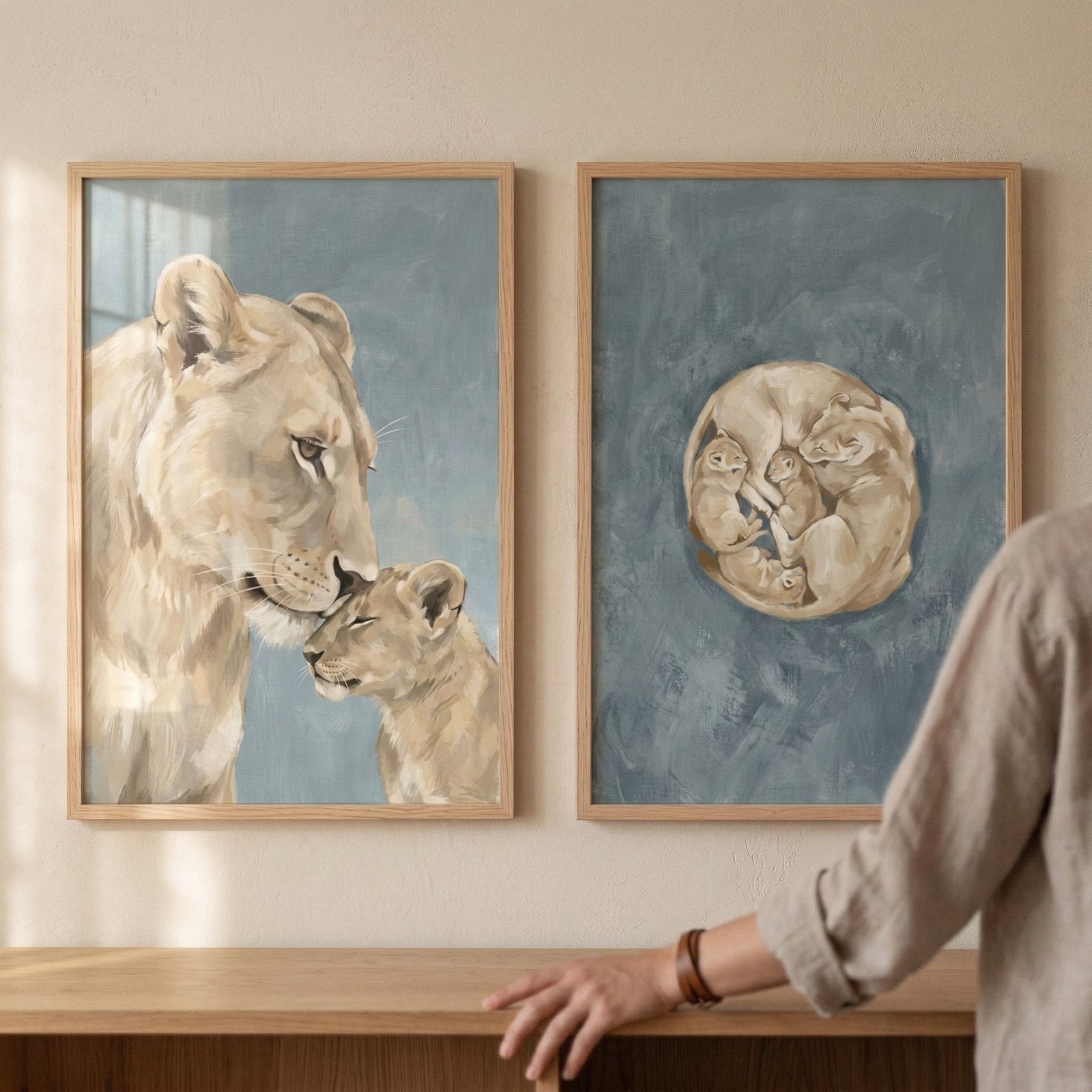 "Two framed illustrations hang side-by-side on a textured, neutral-toned wall above a light wood surface. Both pieces feature a soft, muted color palette with sandy tones for the subjects and a desaturated blue for the background.

The frame on the left showcases a tender scene of a lioness and her cub. The larger lioness is shown from the side, her head bowed as she gently nuzzles the small cub, who has its eyes closed in a peaceful expression. The frame on the right displays a cluster of lion cubs curled 