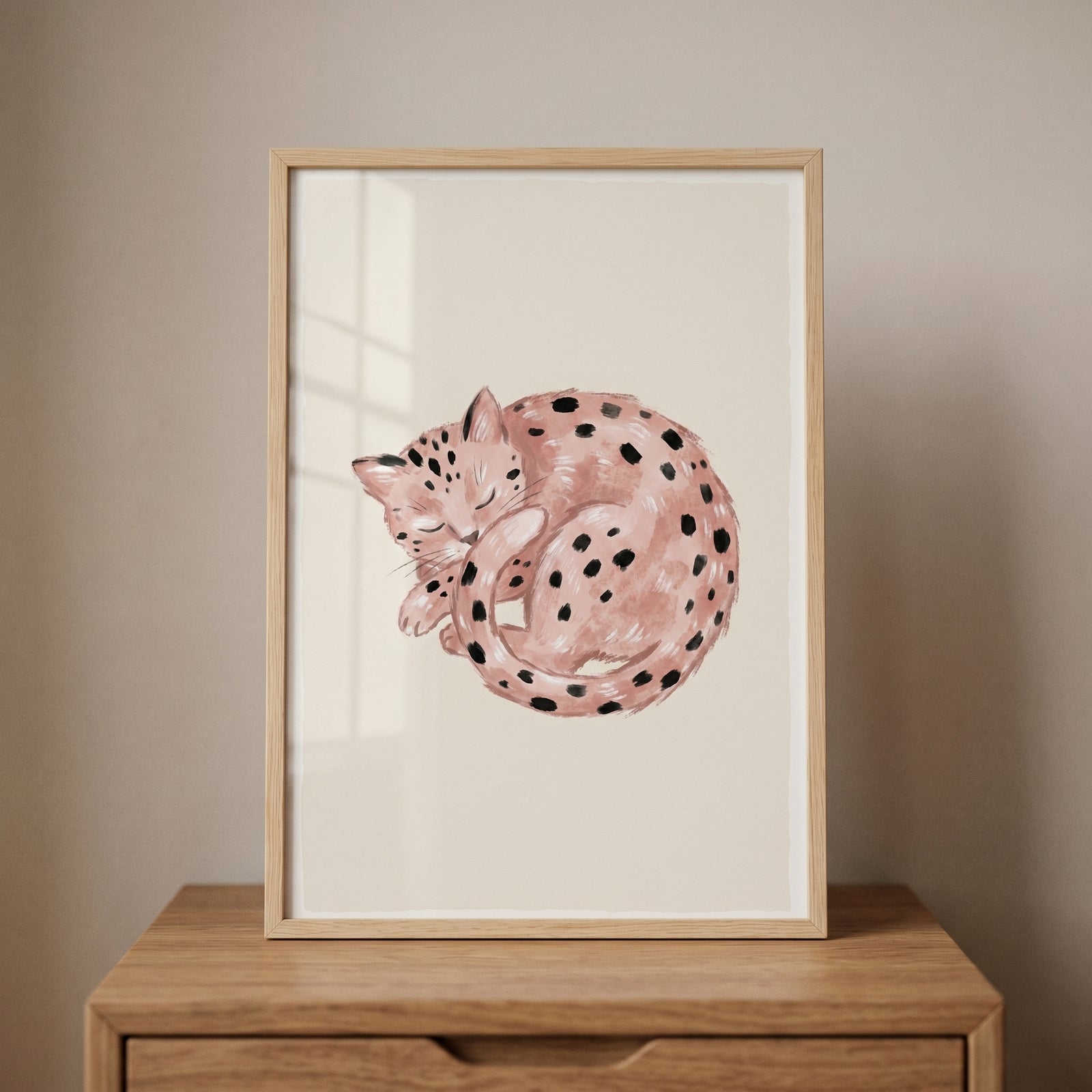 Watercolor pink curled up spotted cat wall art in a light wood frame sits on a wooden dresser against a neutral wall.
