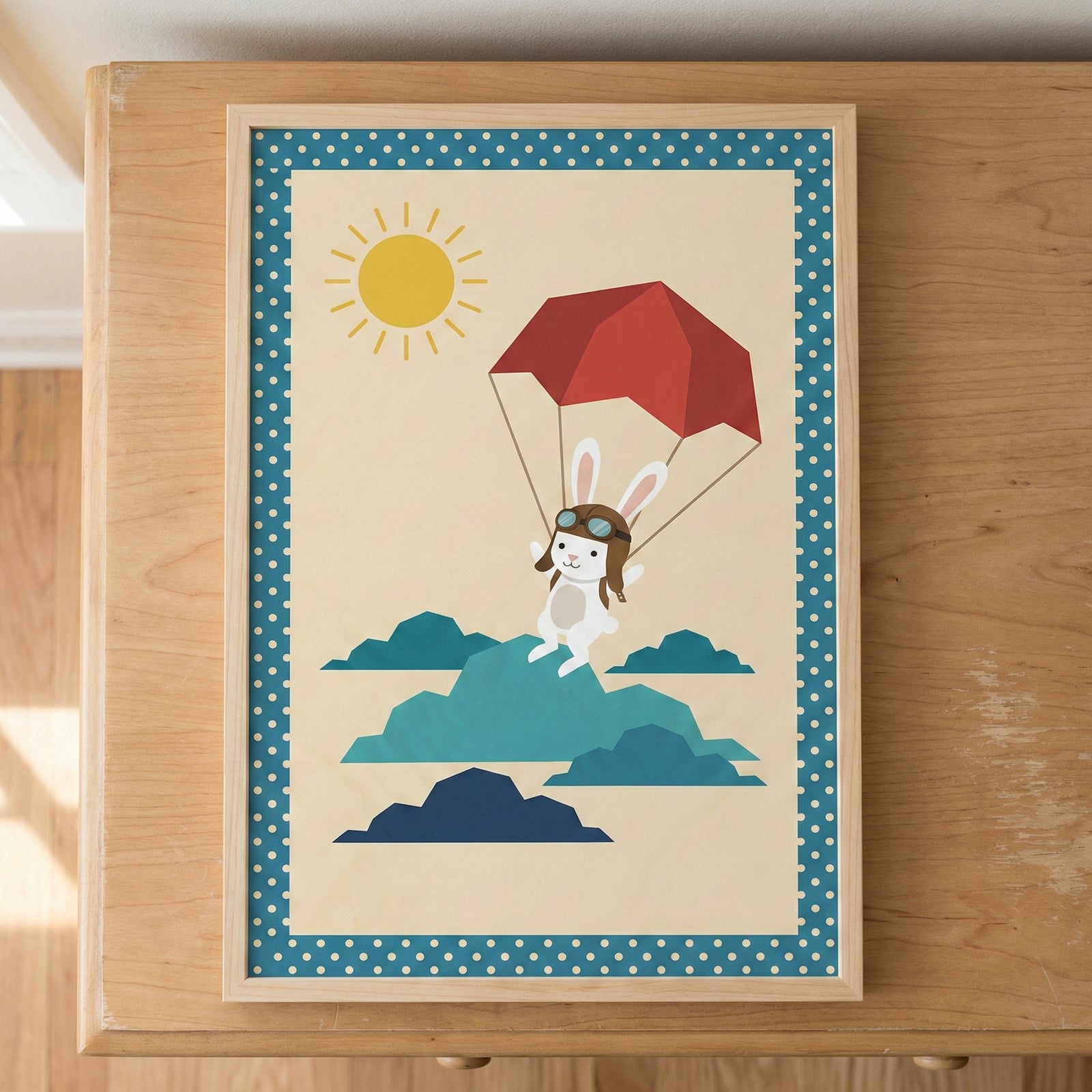 A framed piece of wall art depicting a cute white cartoon rabbit floating in the sky with a red parachute. The rabbit wears brown aviator goggles and a cap. A yellow sun is in the upper left corner, and several stylized blue and teal clouds are at the bottom. The artwork has a blue border with white polka dots and is in a light-colored wooden frame resting on a wooden surface.
