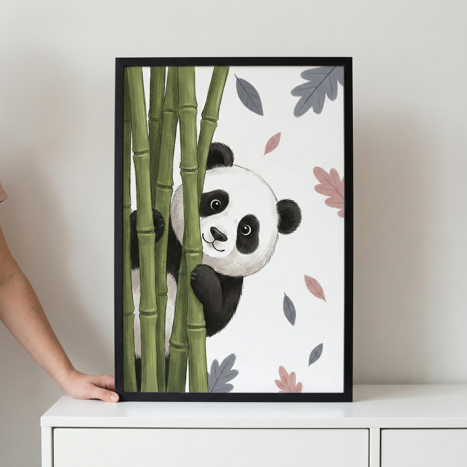 A framed illustration featuring a cute cartoon panda peeking from behind green bamboo stalks. The panda has a friendly expression and is set against a plain white background with scattered, stylized leaves in shades of pink and grey. The artwork is in a thin black frame, resting on a white cabinet, with a person's hand visible on the left side holding the frame.