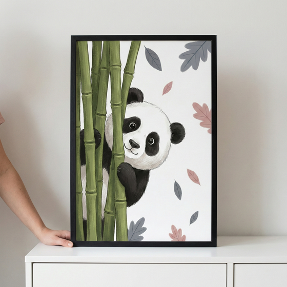 A framed illustration featuring a cute cartoon panda peeking from behind green bamboo stalks. The panda has a friendly expression and is set against a plain white background with scattered, stylized leaves in shades of pink and grey. The artwork is in a thin black frame, resting on a white cabinet, with a person's hand visible on the left side holding the frame.