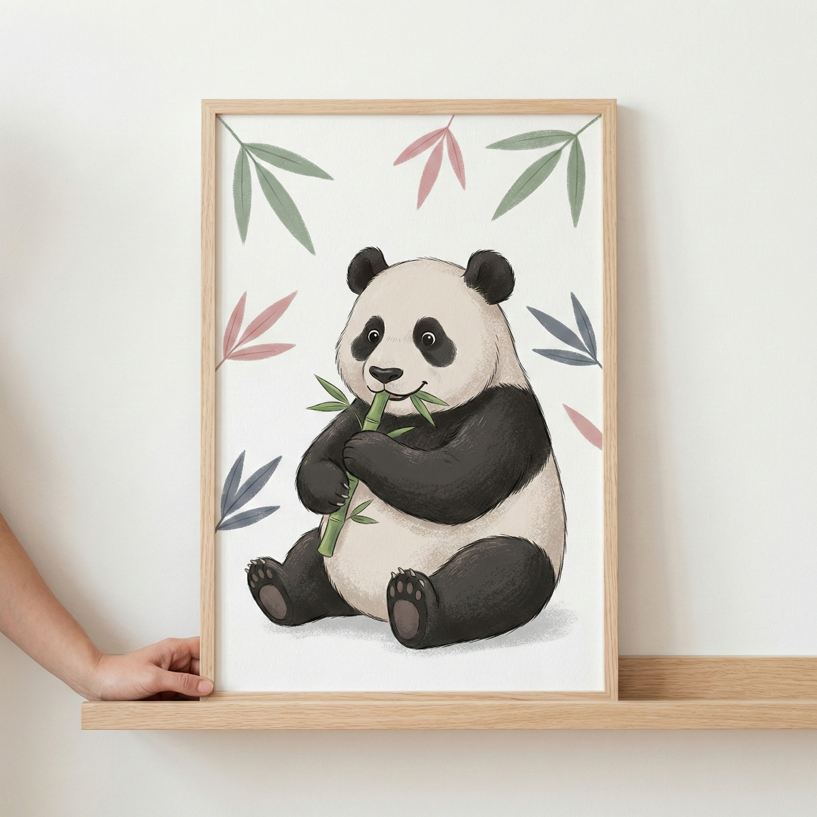 A soft illustration of a giant panda sitting and eating a piece of bamboo is shown in a light wood frame. The panda, with its distinctive black and white markings, holds a green bamboo stalk to its mouth. The background is white, decorated with stylized leaves in muted shades of pink, green, and blue. The framed artwork is being held by a hand and rests on a light-colored wooden shelf against a plain white wall.