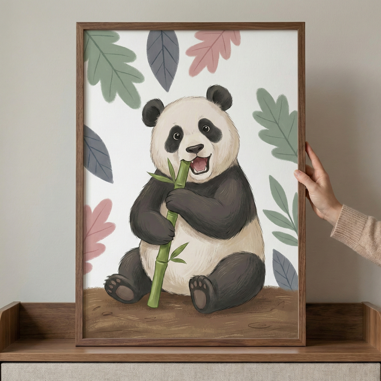 A framed digital illustration features a happy cartoon panda sitting on the ground. The panda is depicted with a cheerful expression, holding and biting into a green bamboo stalk. The background is white, adorned with stylized leaves in shades of muted pink, green, and grey-blue. The artwork is encased in a brown wooden frame, which is held by a hand on the right side. The framed piece rests on a wooden ledge against a neutral-colored wall.