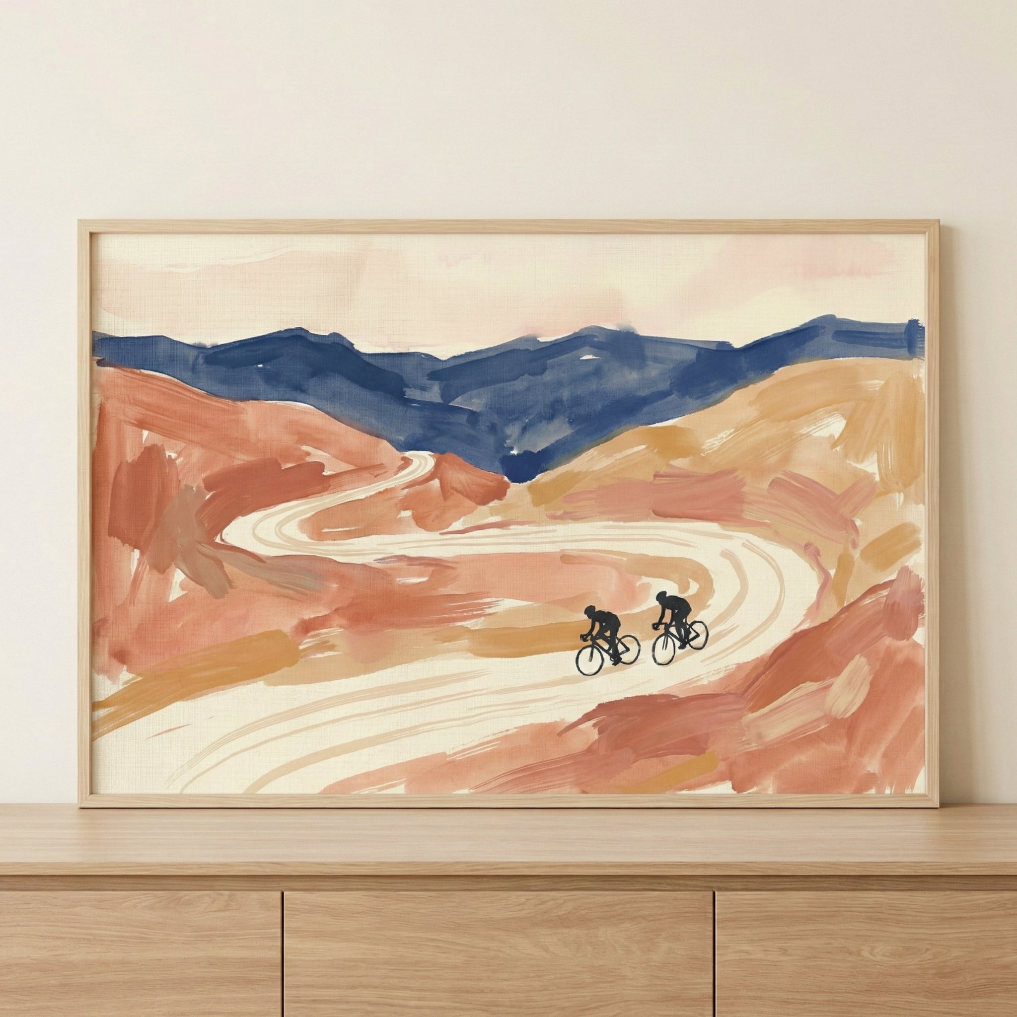 A framed landscape artwork in a loose, expressive style featuring earthy terracotta and peach tones with a dark blue mountain range in the background. A light-colored winding road meanders through the terrain, with two small black silhouettes of cyclists riding along it. The piece is displayed in a light wood frame resting on a wooden surface.