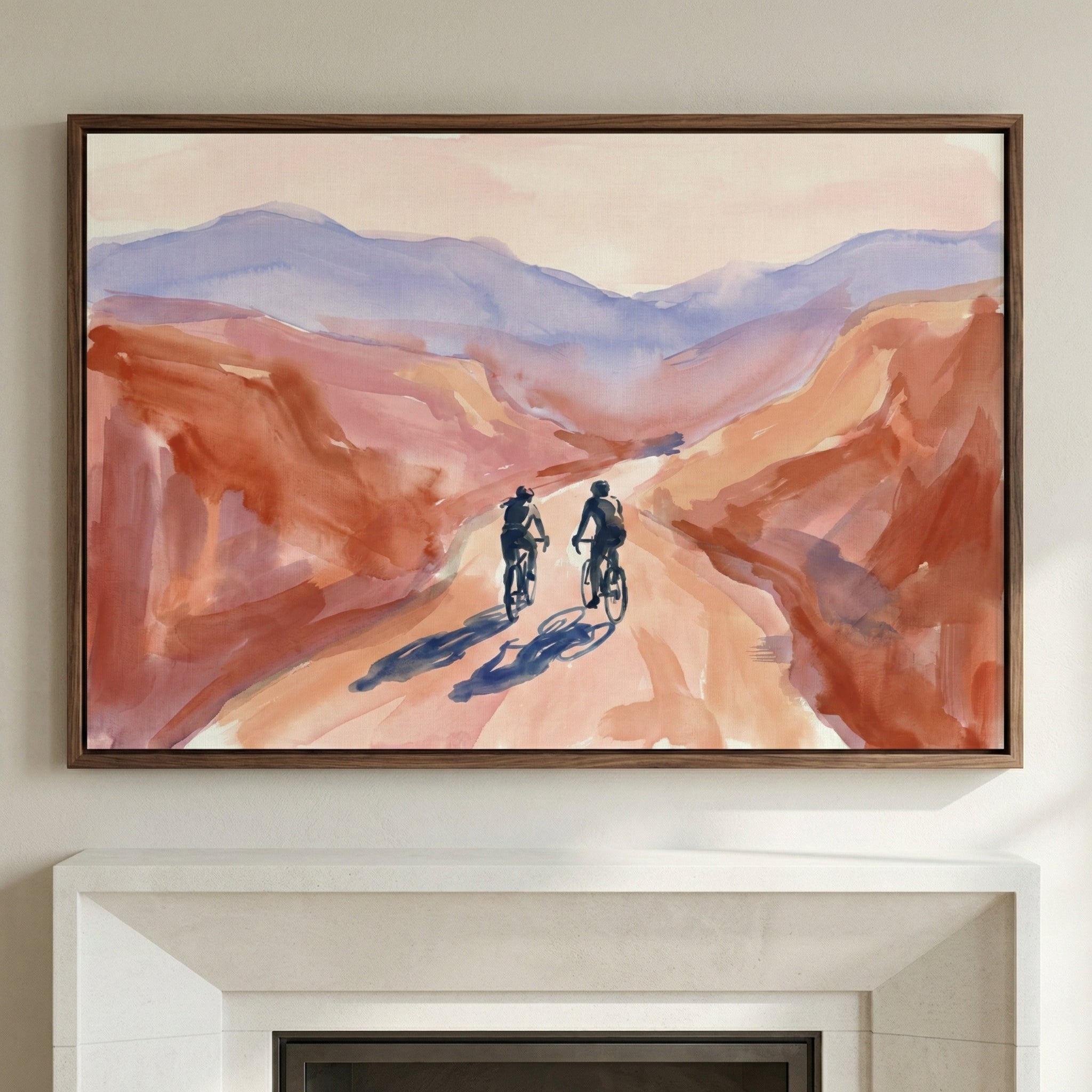 A framed piece of art in a watercolor style hanging above a fireplace mantel. The artwork depicts two people on bicycles riding down a light-colored path through a canyon. The surrounding landscape features warm shades of terracotta and orange, while distant mountains are rendered in soft lavender and blue hues. Long shadows stretch across the trail in front of the cyclists under a pale, warm sky.