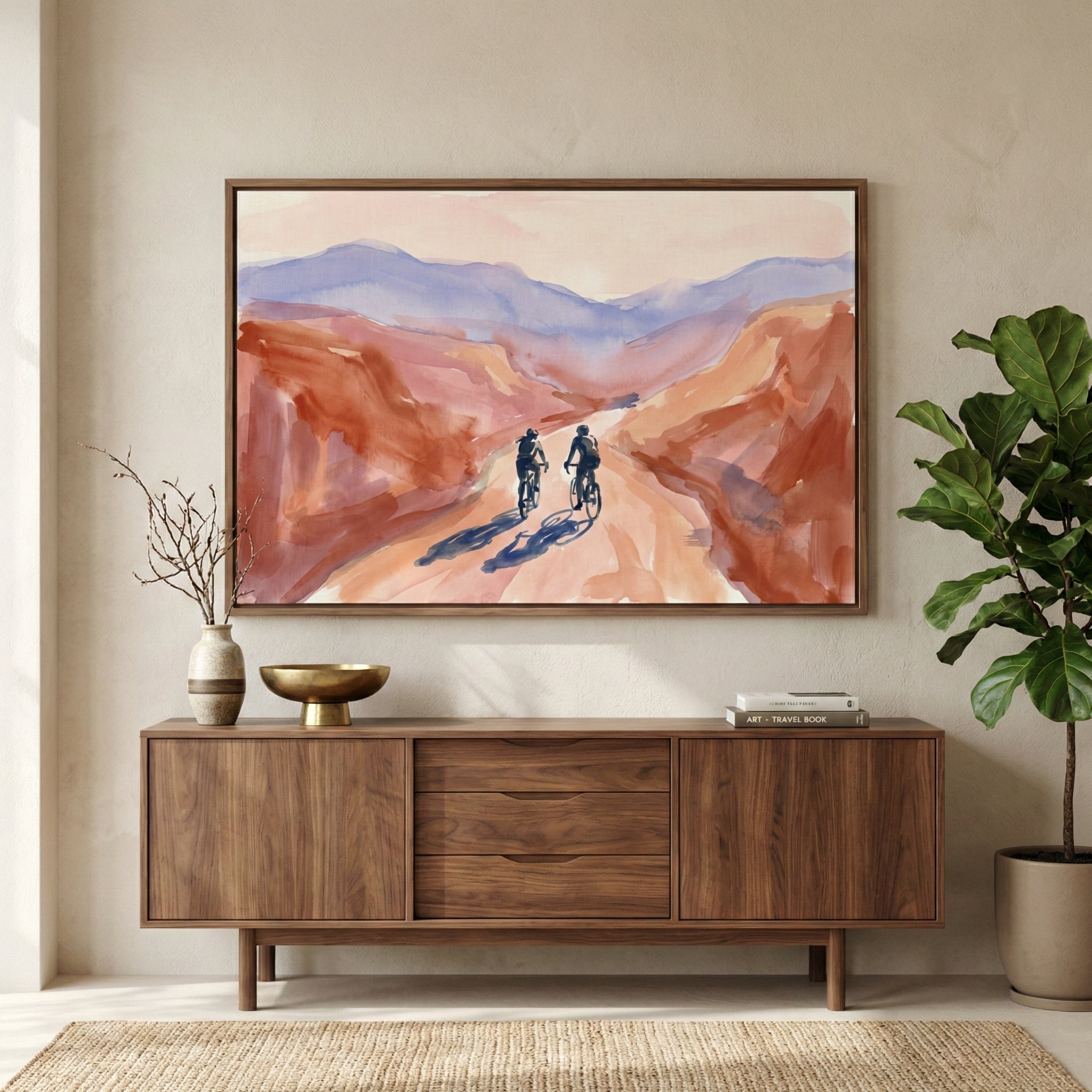 A horizontal framed watercolor-style landscape hangs on a neutral-toned wall above a mid-century modern wooden sideboard. The artwork depicts two individuals riding bicycles on a light-colored path winding through a vast canyon with rolling mountains in the distance, using a color palette of warm terracotta, muted oranges, and soft lavender. On the wooden sideboard sits a small ceramic vase with thin branches, a gold-colored pedestal bowl, and a small stack of books. 