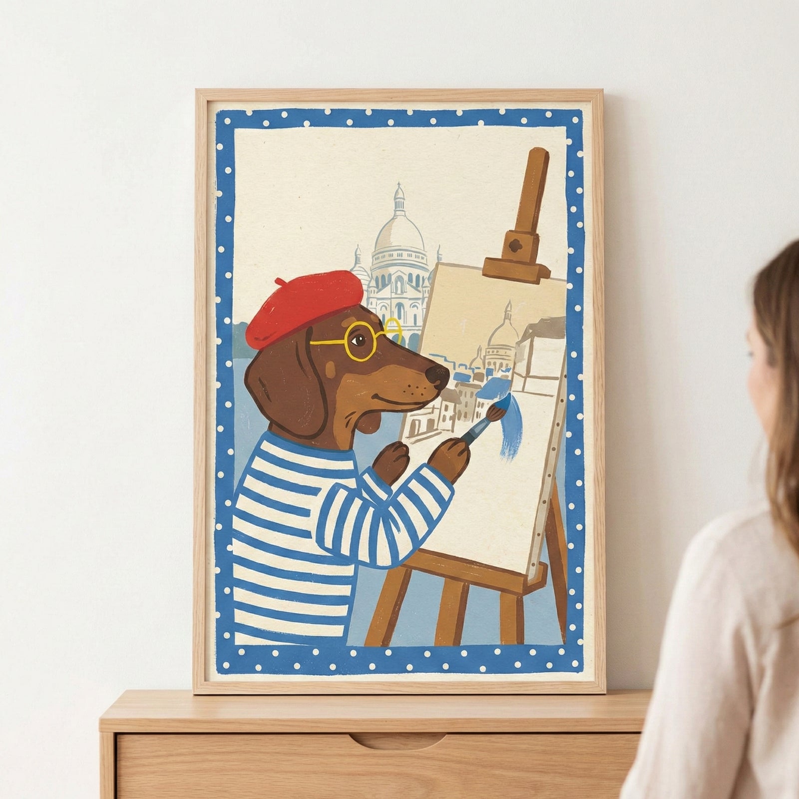 A framed illustration shows a cartoon dachshund dressed in a red beret, yellow glasses, and a blue and white striped shirt. The dog uses a brush to apply blue color to a sketch on an easel that depicts a cityscape with a large white domed building. The image is bordered in blue with white polka dots and sits in a light wood frame on a wooden dresser.
