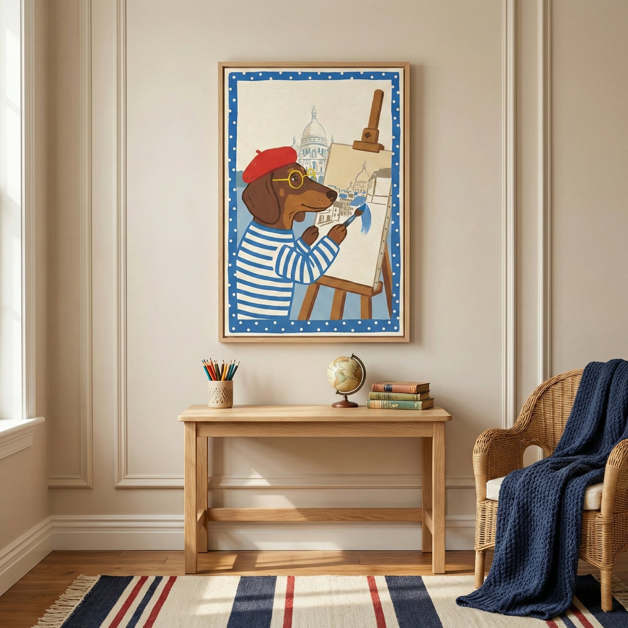 A framed illustration on a neutral wall depicts a dachshund wearing a red beret, yellow glasses, and a blue and white striped shirt, working at an easel with a brush. Below the artwork, a light wood desk holds several books, a small globe, and a cup of colored pencils. To the right, a wicker armchair is draped with a textured navy blue throw. A striped rug in red, white, and blue covers the floor in front of the desk.