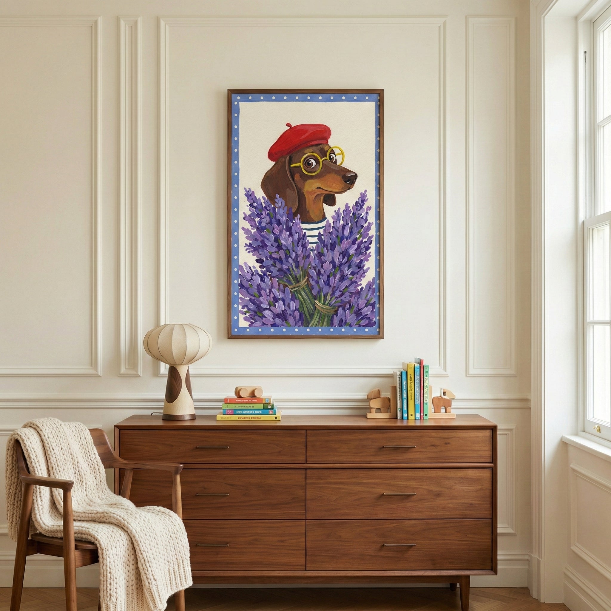A vertical framed illustration hangs centered on a white paneled wall above a mid-century modern six-drawer wooden dresser. The artwork depicts a brown dachshund wearing a red beret and yellow-rimmed glasses, appearing from behind a large bouquet of purple lavender. The image is bordered by a blue frame with white polka dots. On the dresser, there is a rounded beige lamp, a small stack of colorful books, and wooden bookends. To the left, a wooden chair is draped with a textured white throw blanket, and a wh