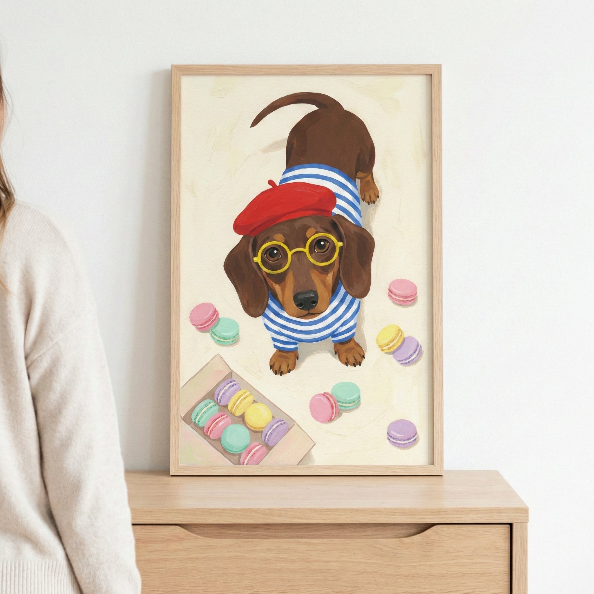 A framed illustration of a brown dachshund dog looking upwards. The dog is wearing a small red beret, round yellow-rimmed glasses, and a blue and white striped sweater. Various colorful macarons in shades of pink, yellow, green, and purple are scattered around the dog on a light background, with some also shown in a small box. The light wood frame is resting on a wooden cabinet next to a person.