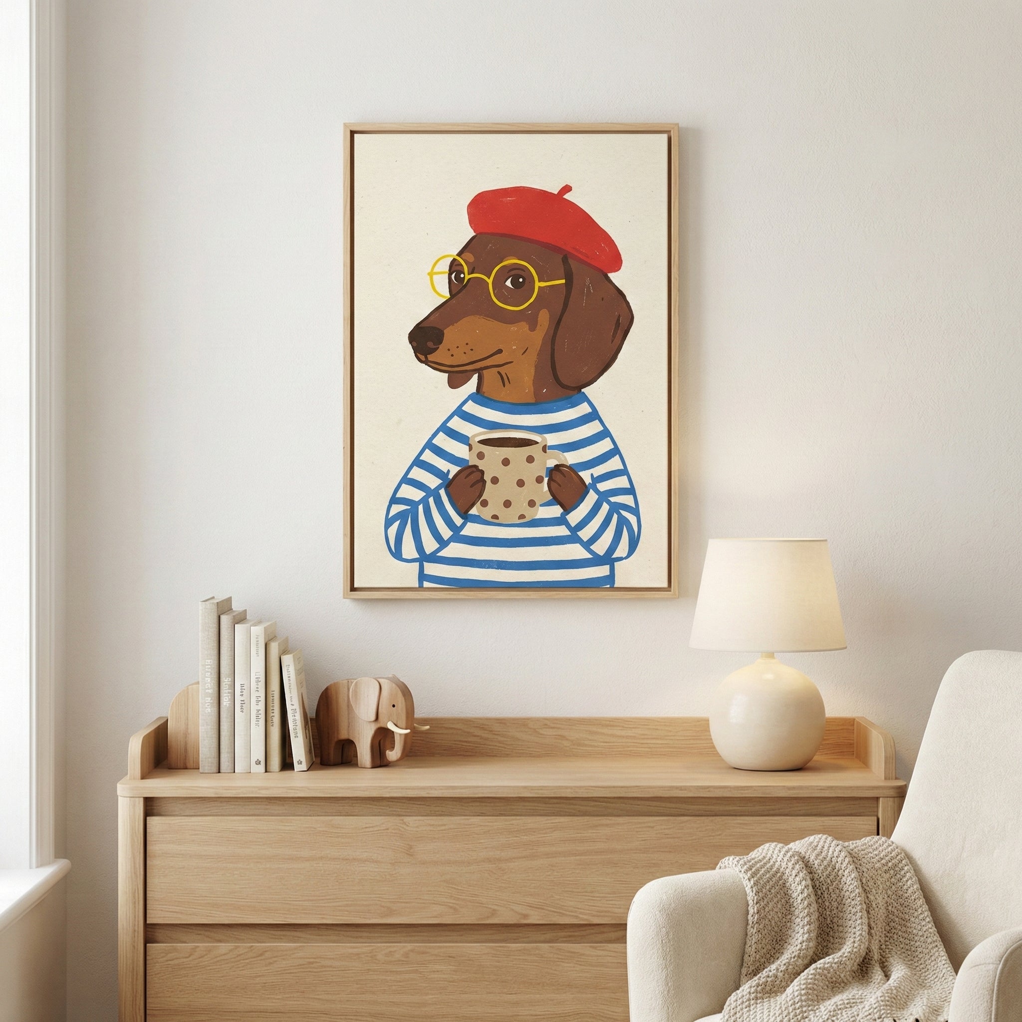 A piece of framed wall art hangs on a light-colored wall above a wooden dresser. The artwork features a whimsical illustration of a brown dachshund dog with human-like features. The dog is wearing a red beret, round yellow glasses, and a blue and white striped shirt, and is holding a beige polka-dotted mug. On the dresser below, there are several books held by a wooden elephant bookend, a small wooden elephant figurine, and a white lamp with a round base. To the right, a white armchair with a textured cream